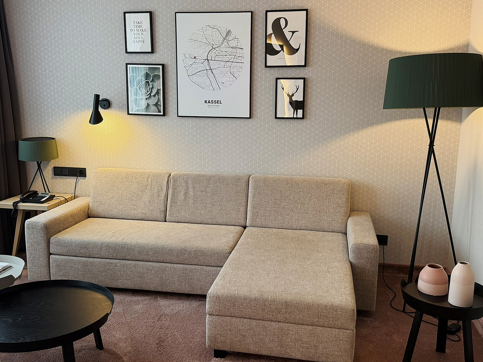 Ein modernes Wohnzimmer mit einem beigen Sofa, das eine L-Form hat. An der Wand hängen mehrere Bilder, darunter ein großer Rahmen mit der Aufschrift "KASSEL". Neben dem Sofa steht ein schwarzer Tisch und eine Lampe mit grünem Lampenschirm.