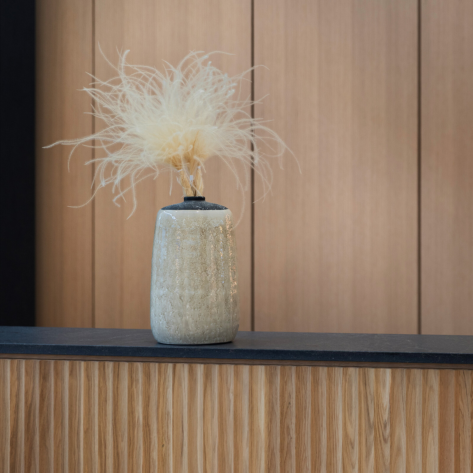 A textured, pale gray vase sits on a dark countertop, holding a floral arrangement with soft, feathery, cream-colored plumes. The background features light wooden panels, enhancing the modern aesthetic of the scene.