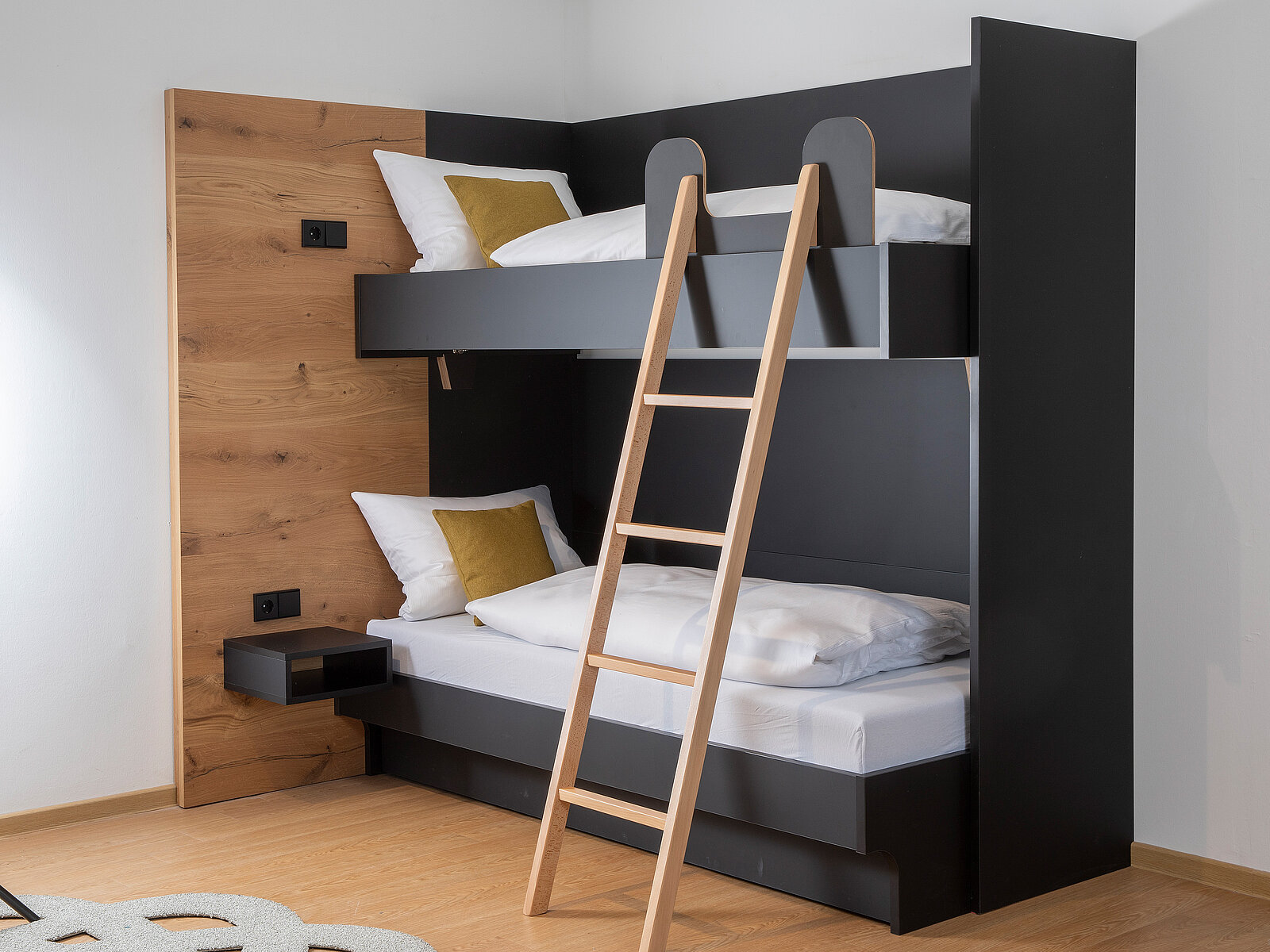 Das Bild zeigt ein modernes, zweigeschossiges Bett aus Holz und dunklem Material. Eine Holzleiter führt zur oberen Matratze. Auf beiden Schlafplätzen liegen weiße Bettdecken und Kissen mit gelben Akzenten. An der Wand sind Steckdosen sichtbar. Der Raum hat einen hellen Bodenbelag.
