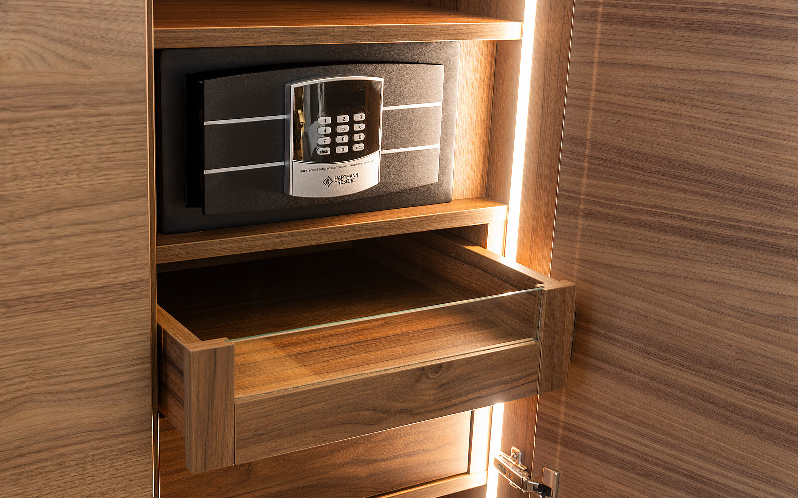 A wooden cabinet features a digital safe with a keypad, positioned in the center. Below the safe, there’s an open drawer with a glass front. The interior of the cabinet is softly illuminated.