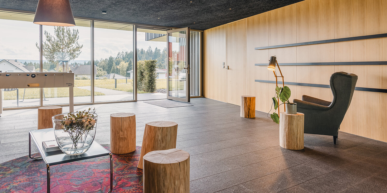 Heller Raum mit großen Glasfenstern, Holzwänden und minimalistischer Einrichtung. Ein schwarzer Sessel neben einer Lampe, Holzhocker und ein Tisch mit Blumen. Im Hintergrund ein Tischfußballtisch. Blick auf einen Garten und bewaldete Landschaft durch die Fenster.