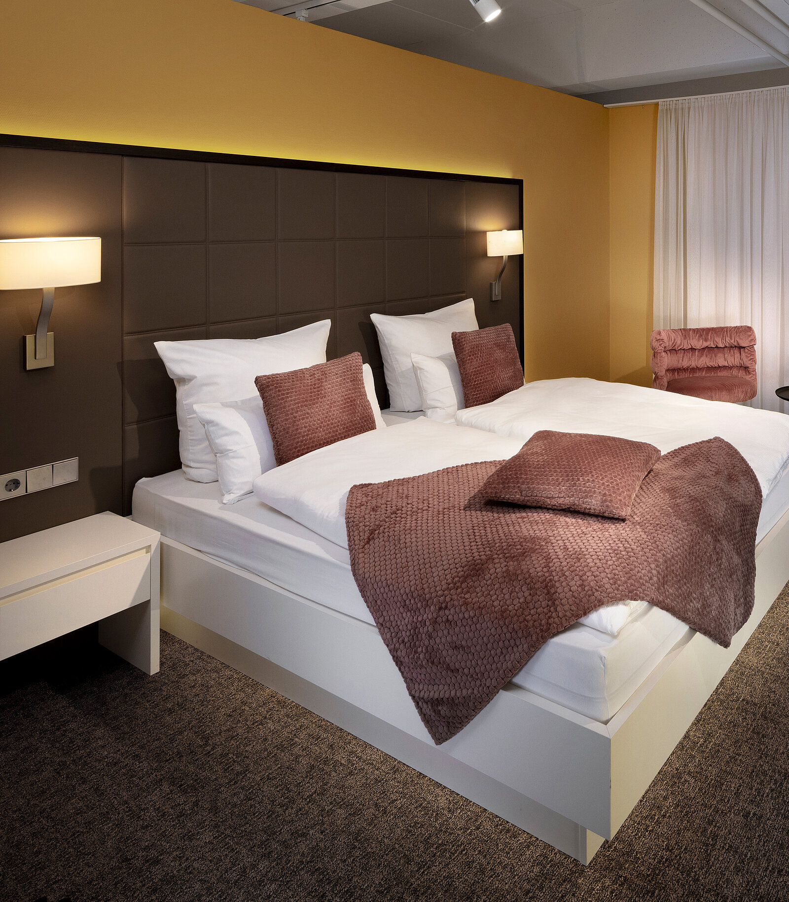 Das Bild zeigt ein modernes Schlafzimmer mit einem großen Doppelbett, umgeben von gemütlichen Kissen und einer Decke in sanften Farben. An der Wand sind zwei Nachtlichter angebracht, und ein bequemer Sessel steht in der Ecke des Raumes. Der Boden ist mit Teppich ausgelegt.