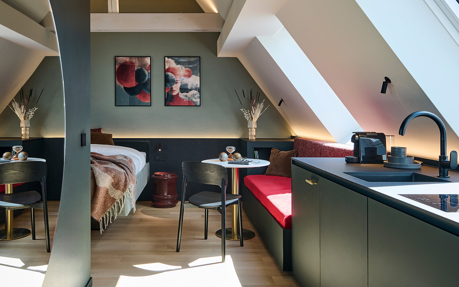 Gemütliches, modern eingerichtetes Dachzimmer mit schrägem Dachfenster. Links ein Bett, in der Mitte ein kleiner Esstisch mit zwei Stühlen, rechts eine Küchenzeile mit Spüle und Kaffeemaschine. An der Wand hängen zwei abstrakte Bilder, darüber Dekovasen mit Trockenblumen.