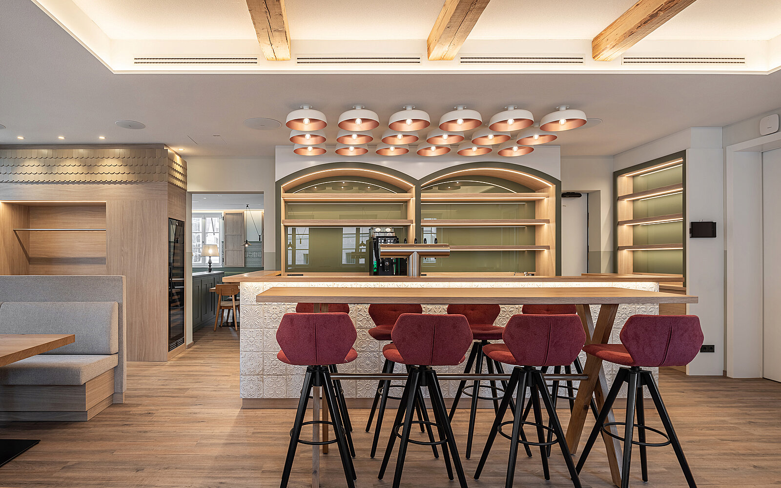 Eine moderne Bar mit einem Holz- und Stein-Mix. Der Bartresen ist hell beleuchtet und von hochmodernen roten Barhockern umgeben. Im Hintergrund sind elegante Regale und eine angenehme Lichtgestaltung zu sehen, die eine einladende Atmosphäre schaffen.