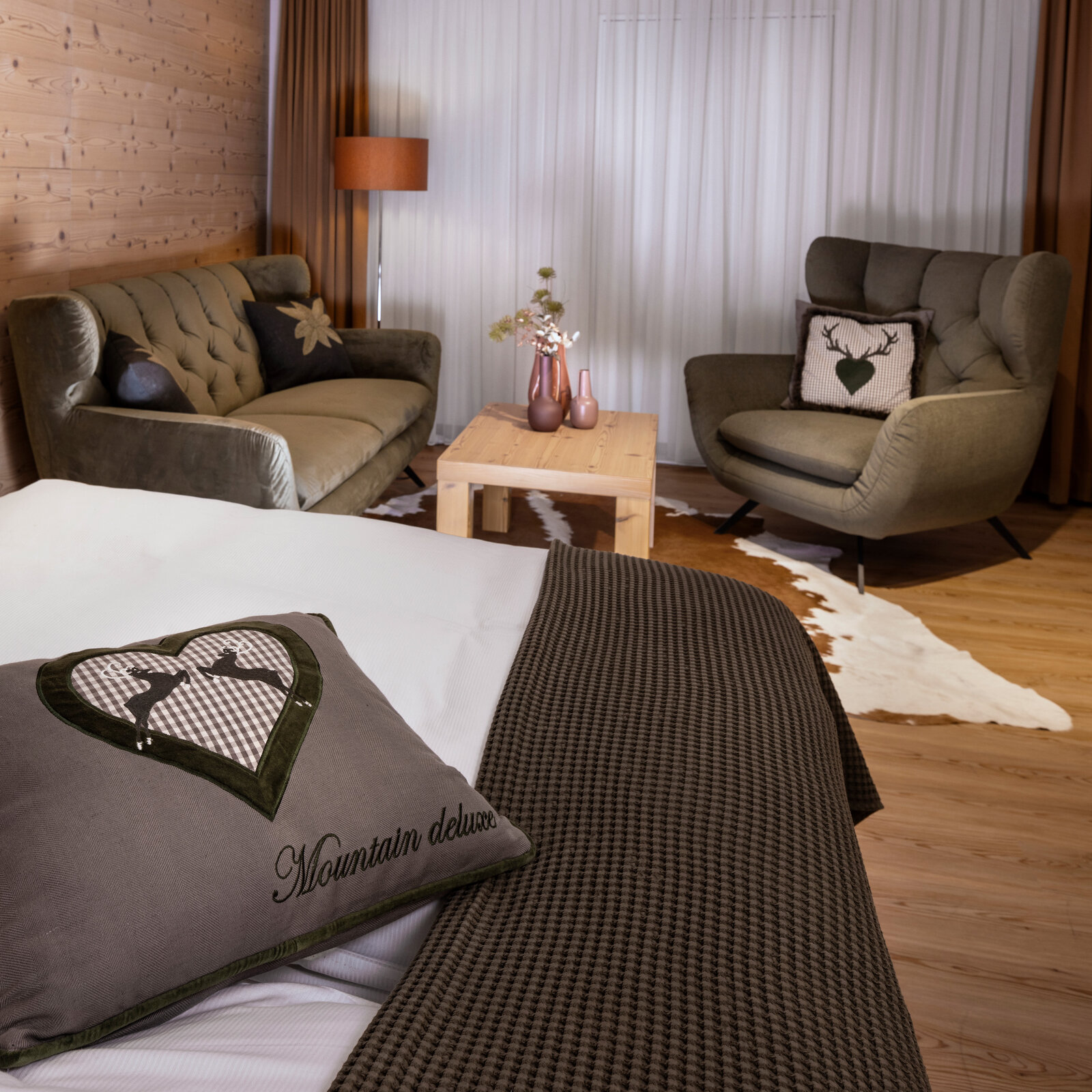 zieflekoch musterzimmer zulux, gemütliches Zimmer mit einem Doppelbett, dekorativen Kissen, einem Sofa, einem Sessel, Holzwänden und einem kleinen Tisch mit Vasen, warme Beleuchtung und Holzfußboden