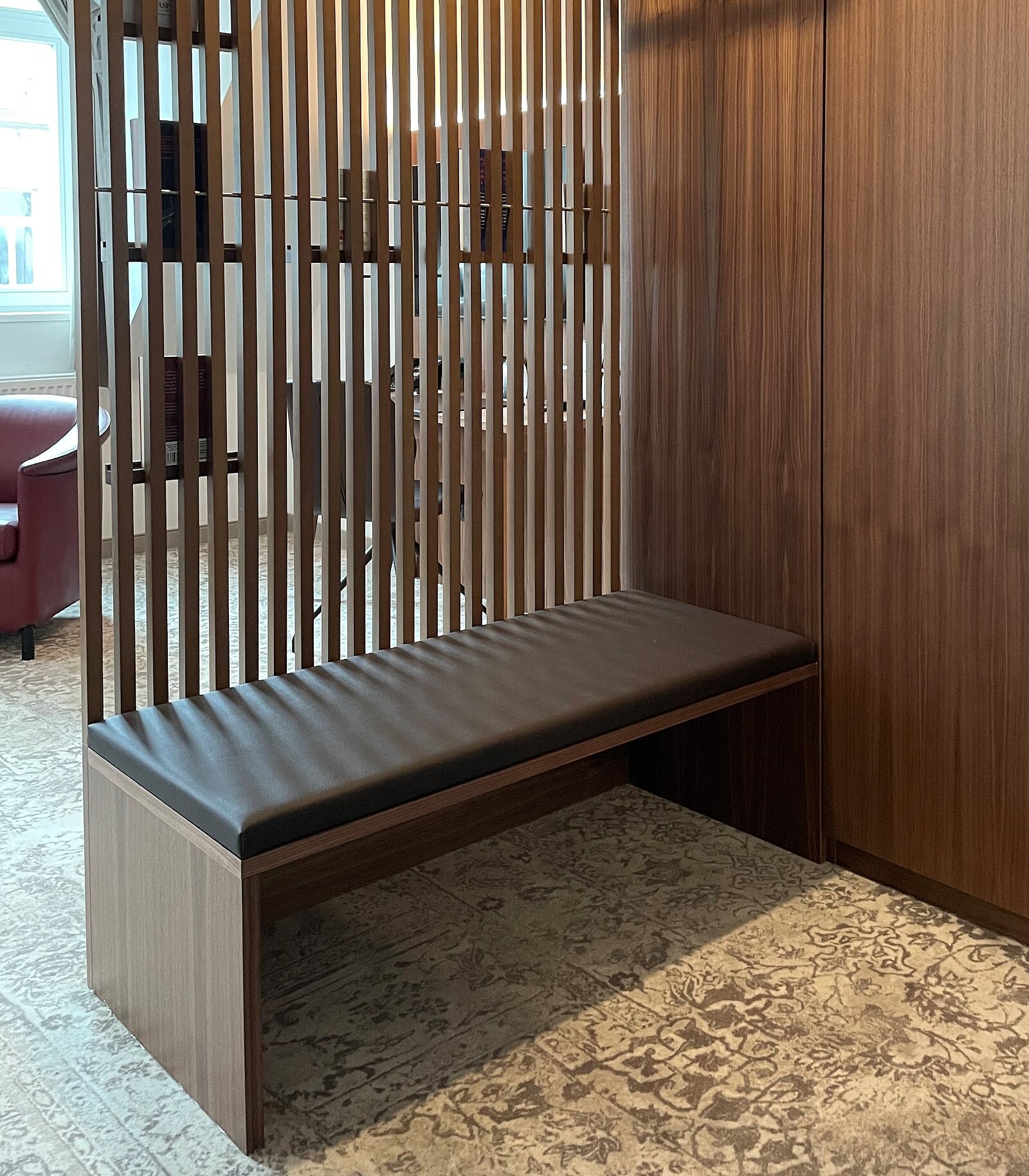 Eine elegante, schlichte Bank aus Holz mit einem gepolsterten schwarzen Sitzpolster steht vor einer Holzlattenwand. Die Umgebung wirkt modern und gepflegt, mit einem Teppich im Hintergrund.