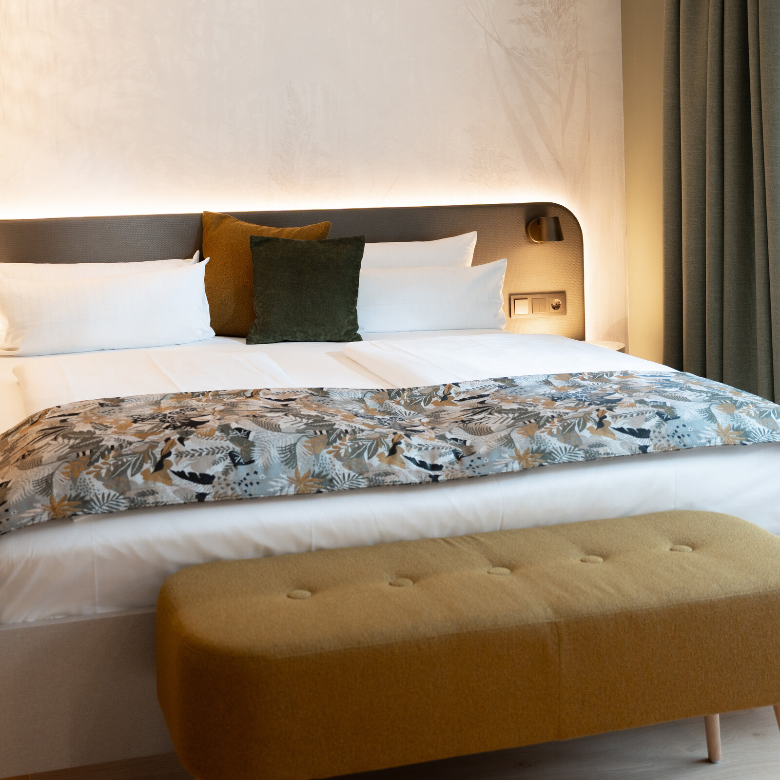 Modern hotel room with a neatly made bed, decorative pillows, a patterned throw, and a mustard-colored bench at the foot.