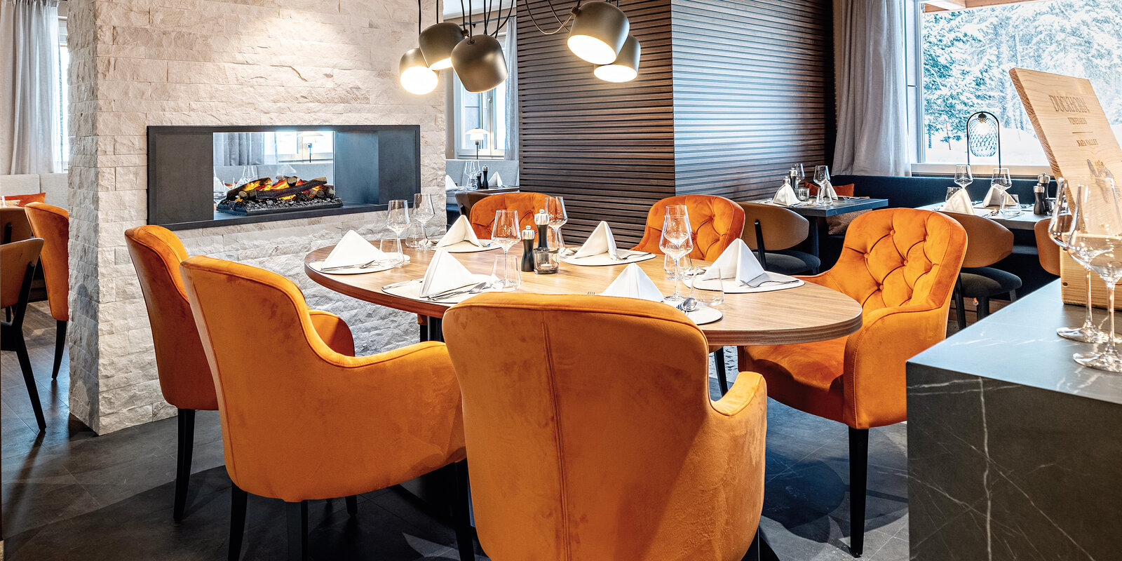 Zieflekoch hotel piz badus Andermatt Schweiz Restaurant Frühstücksraum nussbaum wandbild berge steinwand kamin nussbaum lamellen polstersessel gedeckter Tisch Hängeleuchten winter