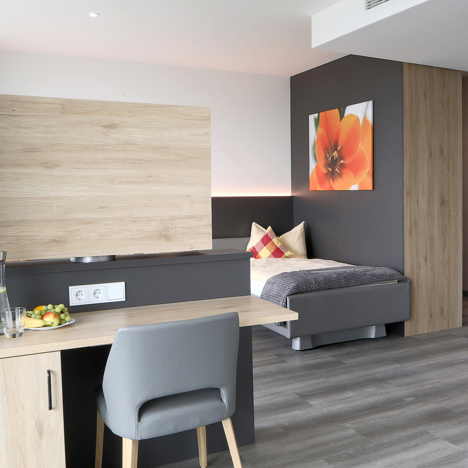Ein modern eingerichtetes Zimmer mit einem kleinen Tisch, einem Stuhl und einer Schale mit Obst. An einer Wand hängt ein Bild einer Blume. In einer Ecke steht ein Bett mit einem Kissen und einer Decke in sanften Farben. Der Boden ist mit Holzoptik ausgelegt.