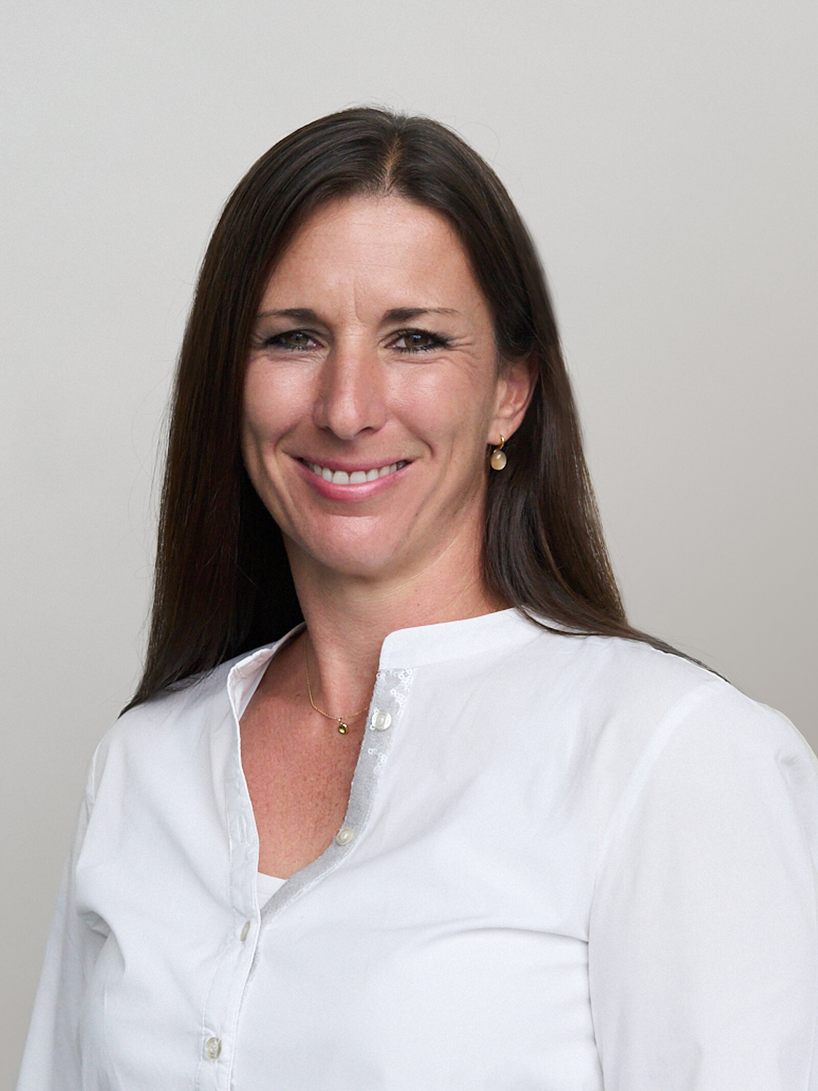 A woman with long, dark hair smiles confidently at the camera. She is wearing a white blouse and a delicate necklace, set against a neutral background. Her expression conveys warmth and approachability.