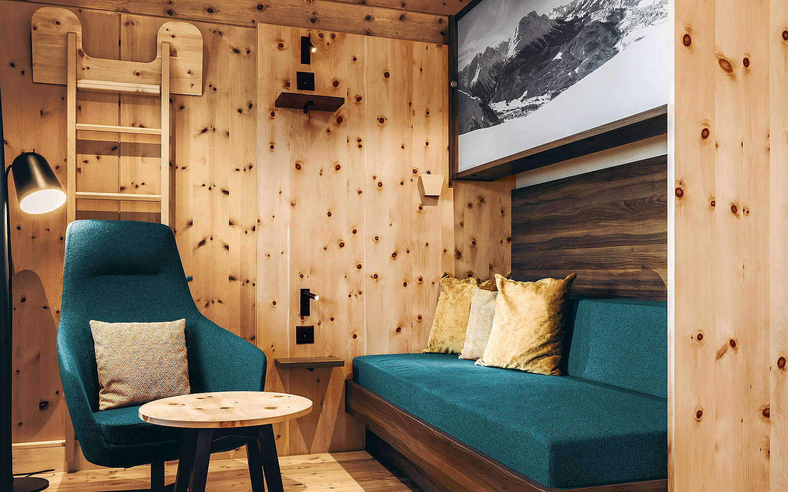 A cozy wooden interior features a teal chair with a cushion and a small round table. A comfortable sofa with decorative pillows sits against the wall, and a ladder leads to a higher level. A framed black-and-white mountain photo is displayed above the sofa.