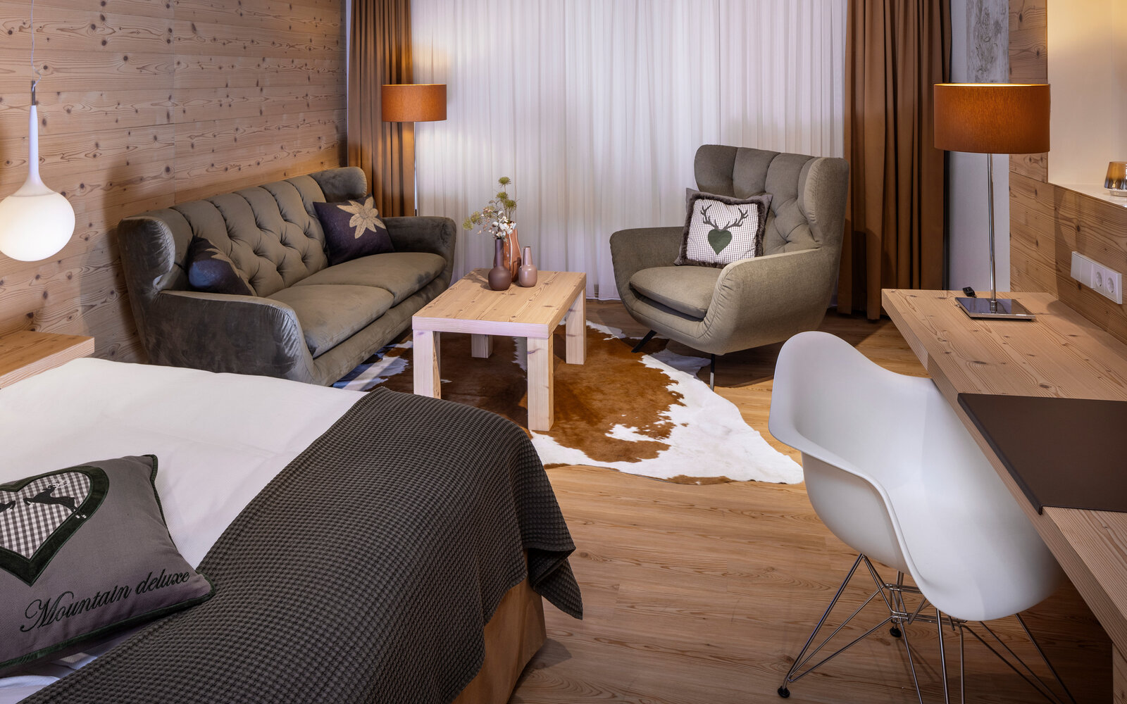 zieflekoch musterzimmer zulux, gemütliches Zimmer mit einem Doppelbett, dekorativen Kissen, einem Sofa, einem Sessel, Holzwänden und einem kleinen Tisch mit Vasen, warme Beleuchtung und Holzfußboden