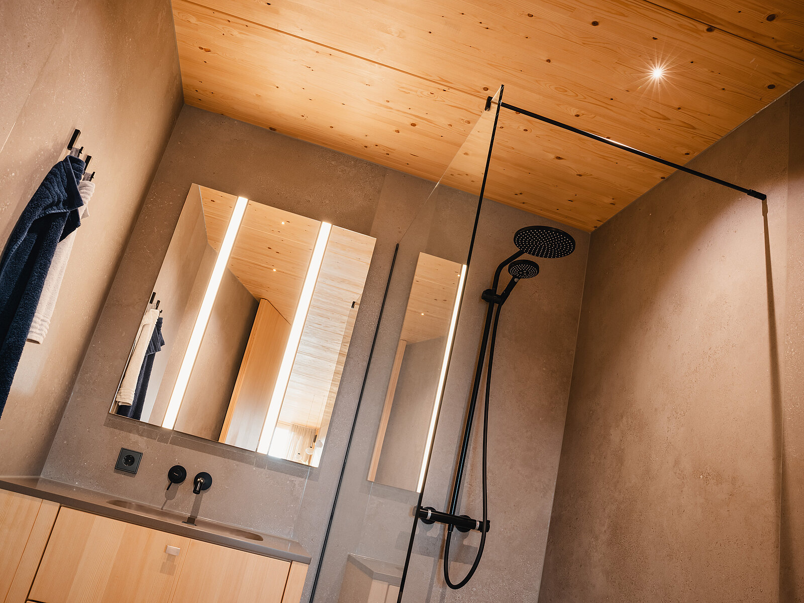 Modernes Badezimmer mit Holzdecke, Spiegeln über einem Waschbecken und einer offenen, begehbaren Dusche mit schwarzer Armatur. Die Wände sind in Betonoptik gehalten, was einen minimalistischen und stilvollen Eindruck vermittelt.