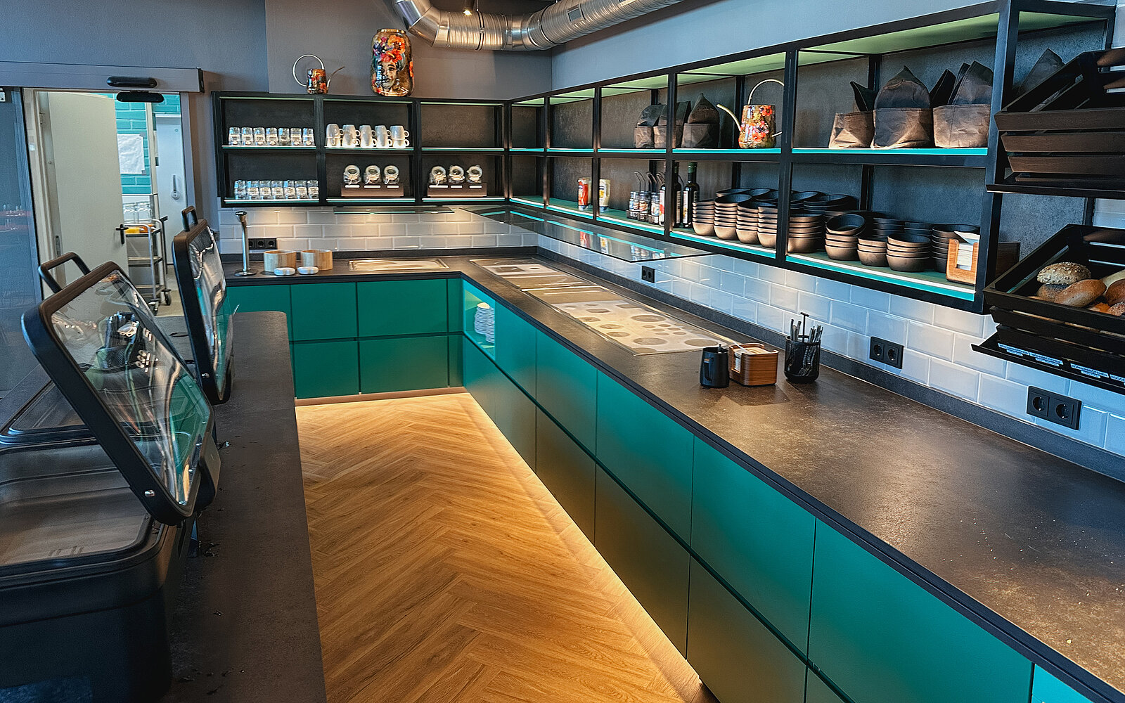 Geschäftsinterieur mit moderner, minimalistischer Kücheneinrichtung. Dunkelgrüne Schränke und dunkle Arbeitsplatten. Freiliegende Regale zeigen Tassen, Schüsseln und Kannen. Parkettboden vervollständigt das Design.