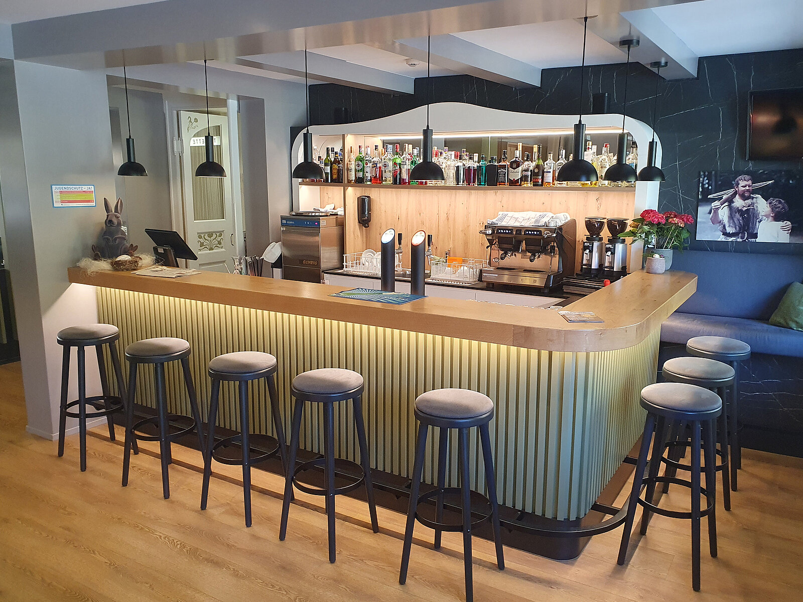 Die Abbildung zeigt eine moderne Bar mit einer geschwungenen Holztheke, die von Senklampen beleuchtet wird. Auf der Theke befinden sich Getränke und eine Kaffeemaschine. Es stehen Stühle in einer Reihe vor der Bar, und im Hintergrund sieht man eine gemütliche Lounge-Ecke.