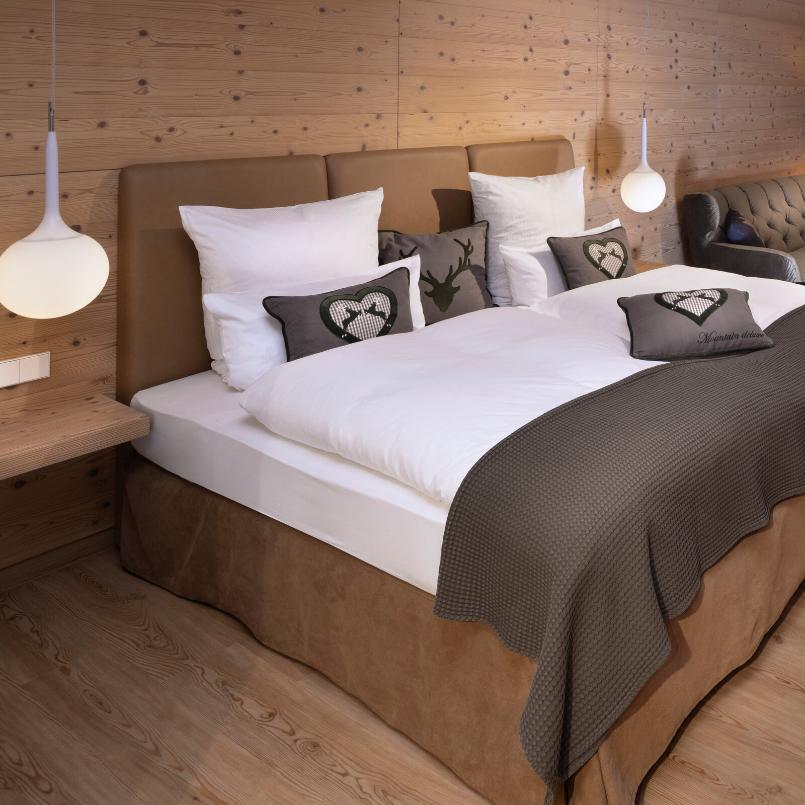 zieflekoch musterzimmer zulux, gemütliches Zimmer mit einem Doppelbett, dekorativen Kissen, einem Sofa, einem Sessel, Holzwänden und einem kleinen Tisch mit Vasen, warme Beleuchtung und Holzfußboden