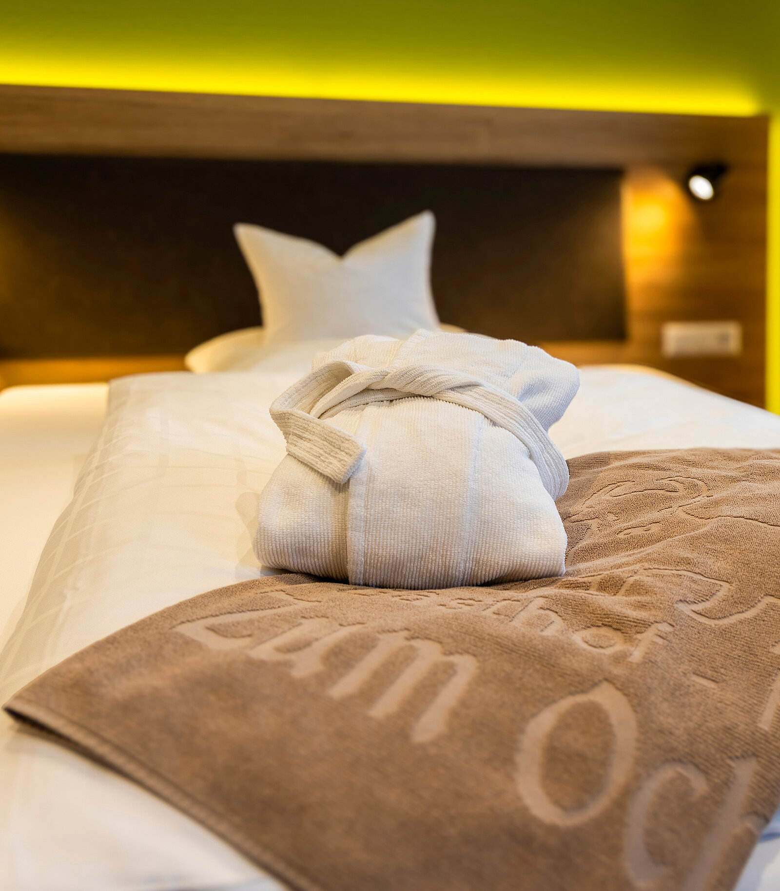 Ein Bett in einem Hotelzimmer mit einem kunstvoll gefalteten Bademantel und einem beigen Handtuch auf der Decke. Das Kopfteil ist aus Holz mit einer indirekten Beleuchtung darüber.