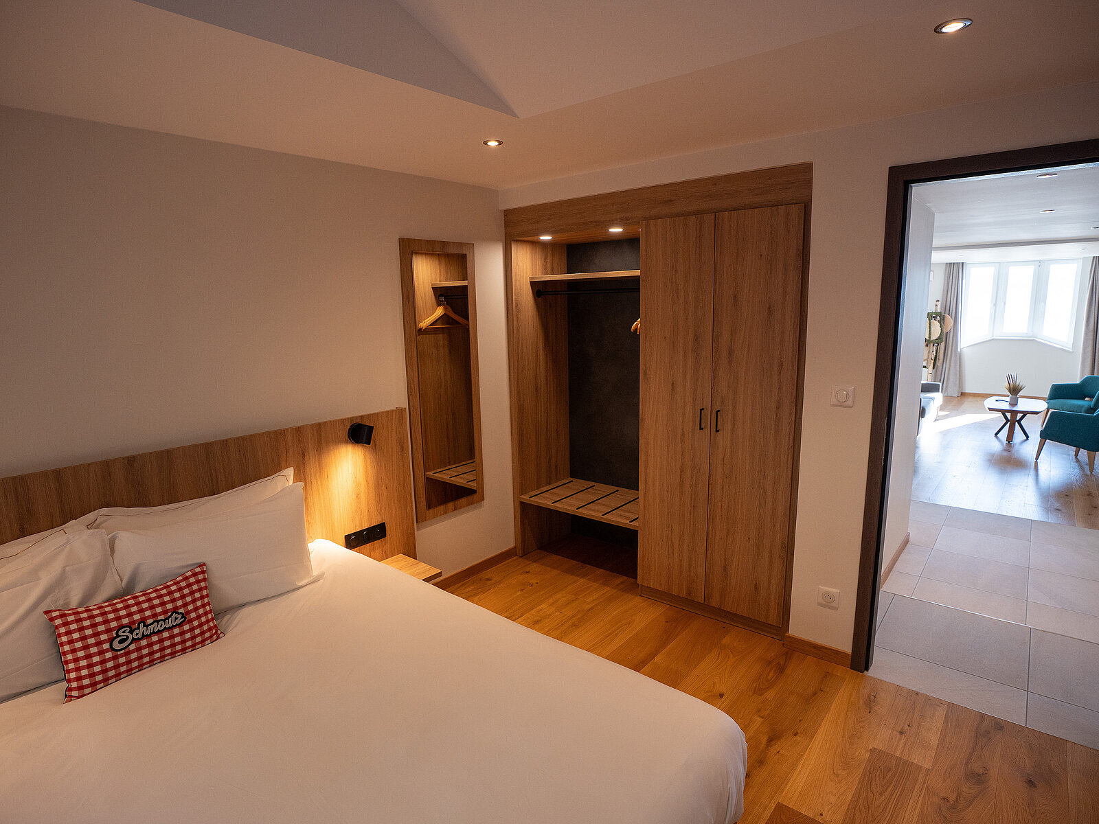 Ein modernes Schlafzimmer mit einem großen Bett, einem roten Zierkissen und einem Holzkleiderschrank mit Spiegel. Der Raum hat Holzböden und eine offene Verbindung zu einem hellen Wohnzimmer mit blauen Möbeln.