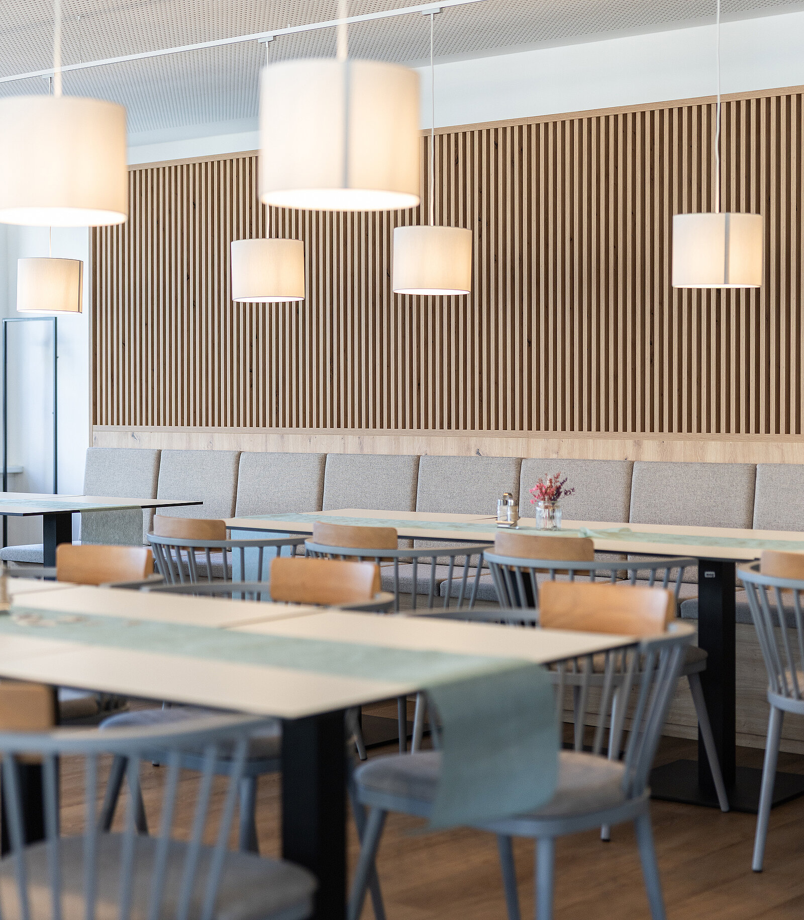 Ein modern eingerichtetes Restaurant mit Holztischen, bequemen Stühlen und mehreren hängenden Deckenlampen. Eine lange Bank steht an der Wand mit senkrechter Holzverkleidung, darauf befinden sich Dekorationen und Blumen. Minimalistisches, warmes Ambiente.