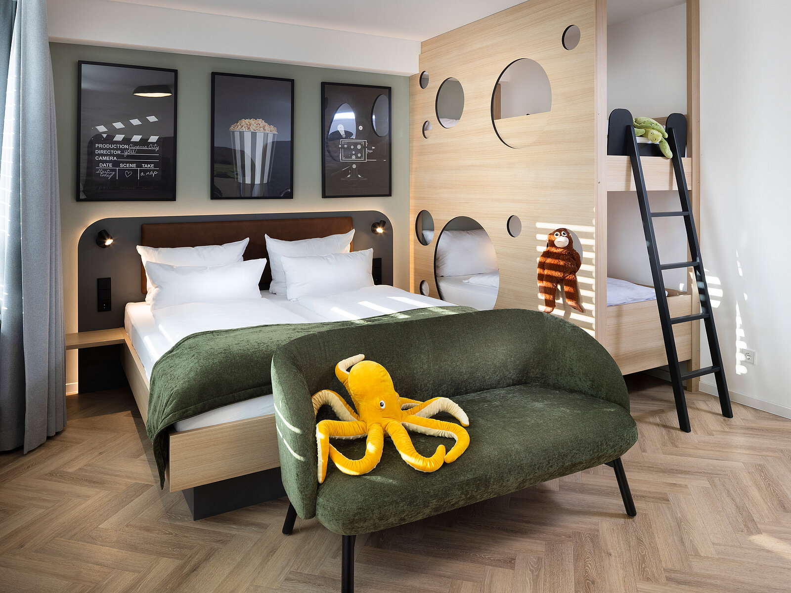 A modern hotel room features a large bed with white linens, a cozy green chair, and playful decor including a stuffed octopus. The room has a unique bunk bed structure with circular cutouts and artwork on the walls depicting popcorn and playful themes.
