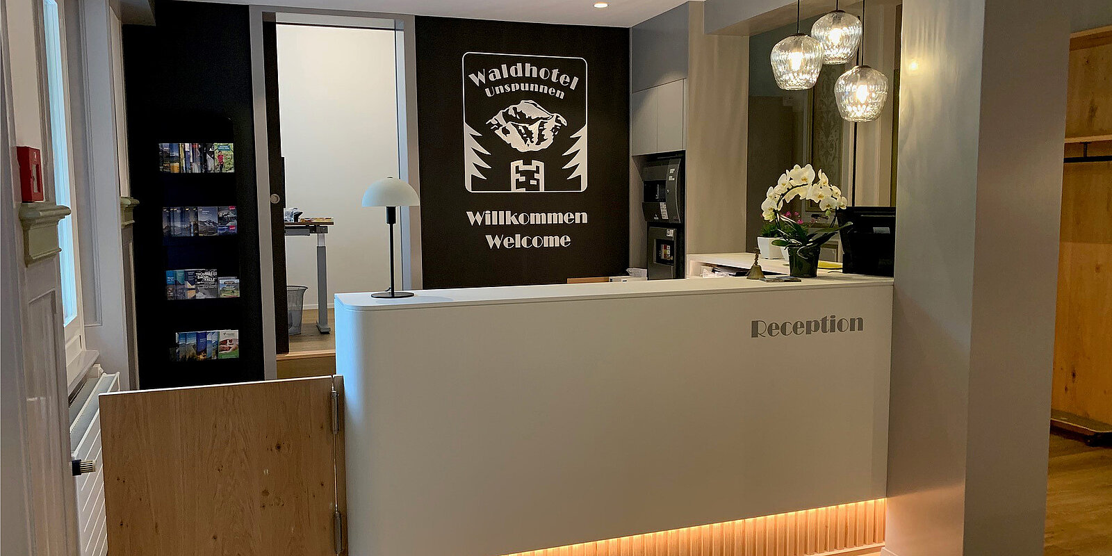 An der Rezeption eines Hotels mit modernem Design steht ein weißer Empfangstresen. Auf dem Tresen befinden sich eine Lampe und eine Blumenvase. Im Hintergrund hängt ein Schild mit der Aufschrift "Willkommen" in Deutsch und Englisch. Hinter der Rezeption sind Informationsmaterialien sichtbar.