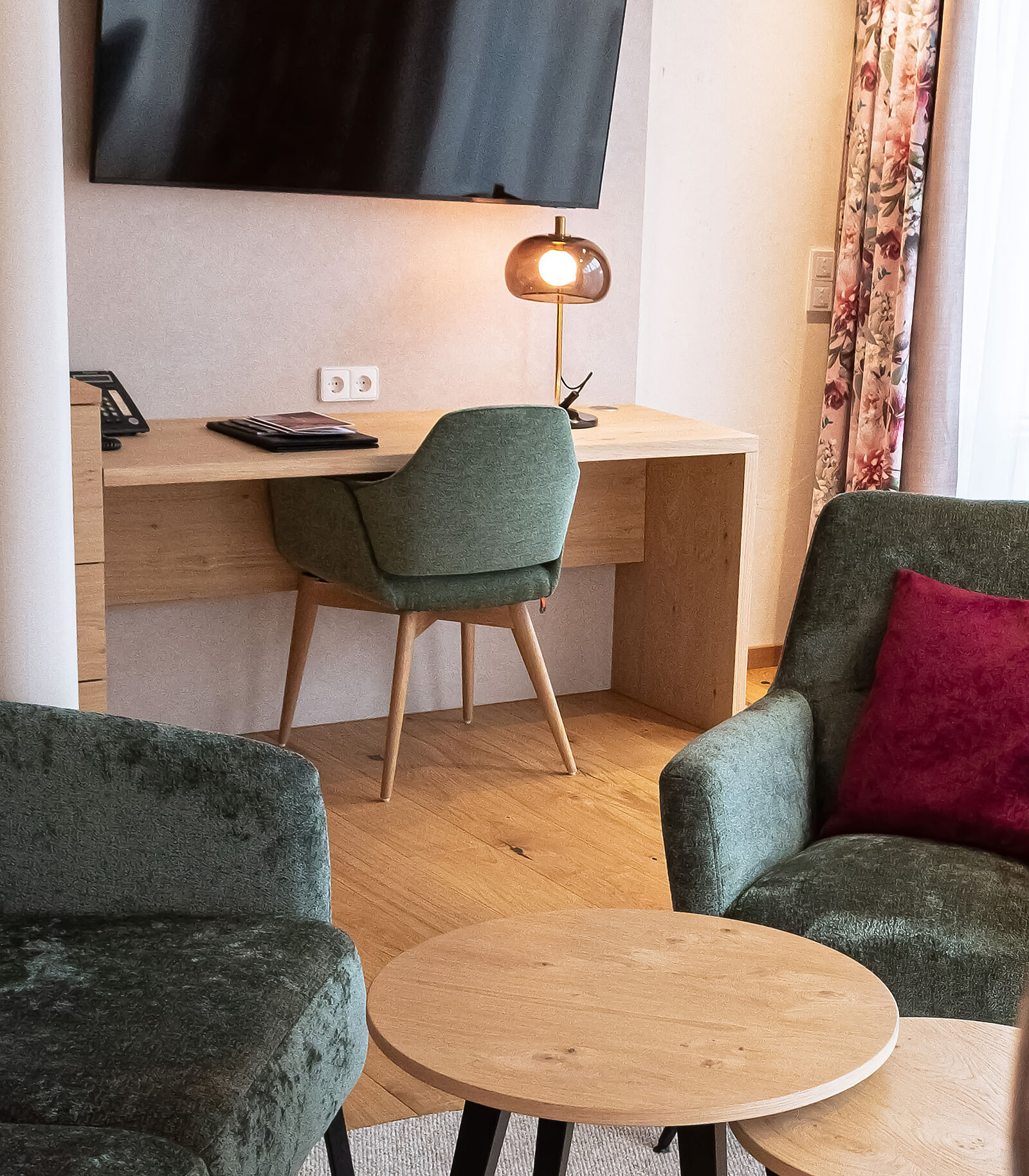 Ein gemütliches Zimmer mit einem Holztisch, einem grünen Stuhl und einer Tischlampe im Hintergrund. Zwei grüne Sessel mit roten Kissen stehen um einen kleinen runden Holztisch. Ein Flachbildfernseher hängt an der Wand.