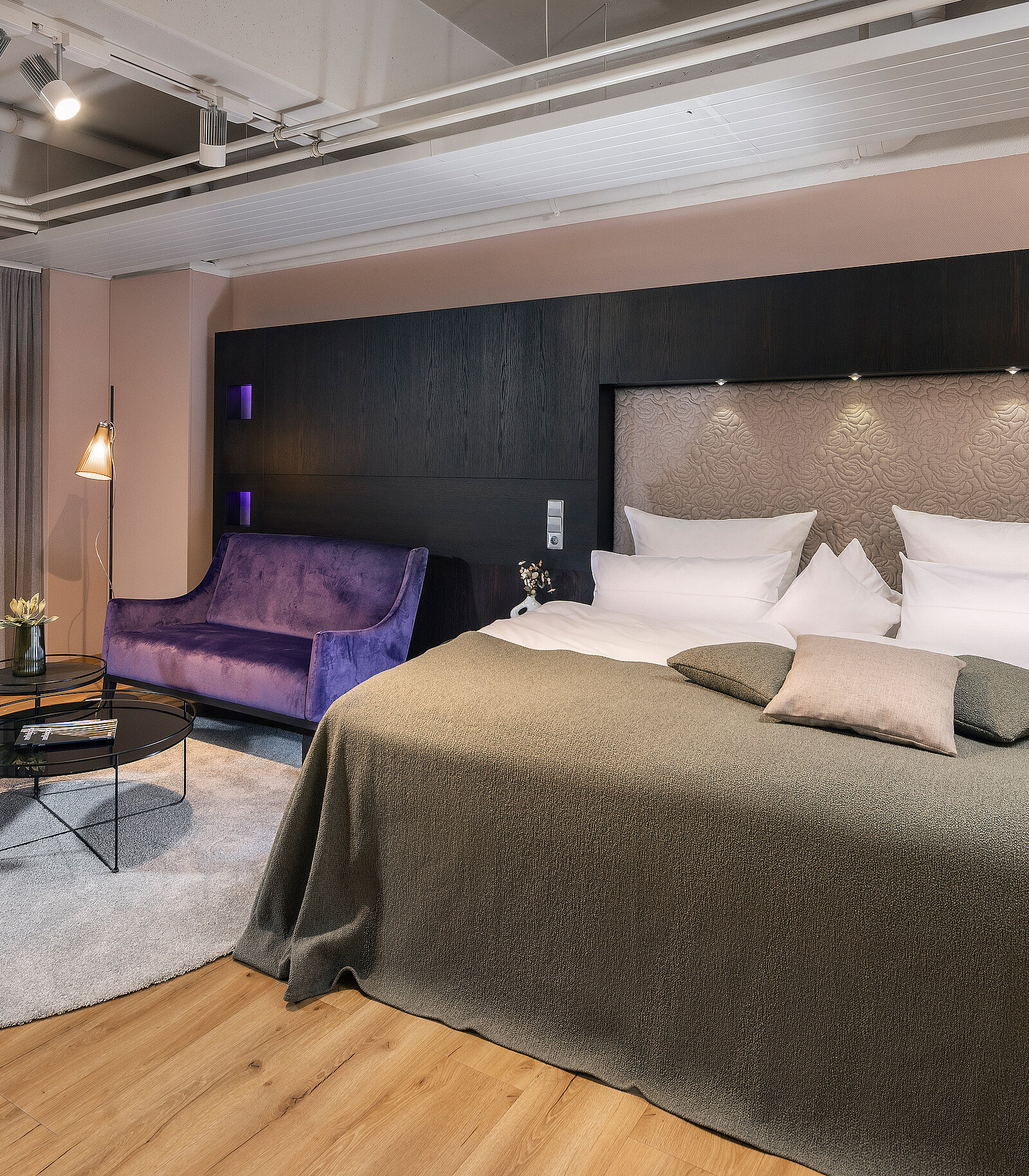 Das Bild zeigt ein modernes Hotelzimmer mit einem großen Bett, das mit einem grauen Überwurf bedeckt ist. Angrenzend befindet sich eine lila Couch und ein runder Kaffeetisch. Der Boden ist aus Holz und der Raum ist in sanften Farben gestaltet.