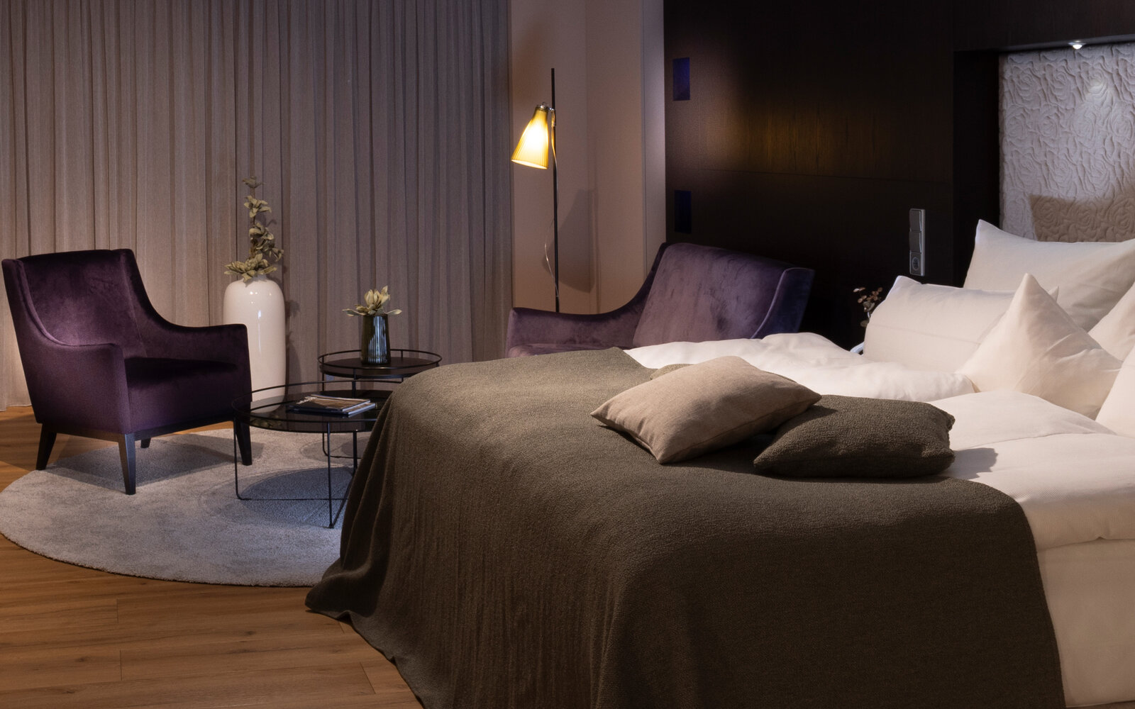 Cozy bedroom with a large bed, purple armchairs, a small round table, and soft lighting. Neutral tones and wooden flooring enhance warmth.