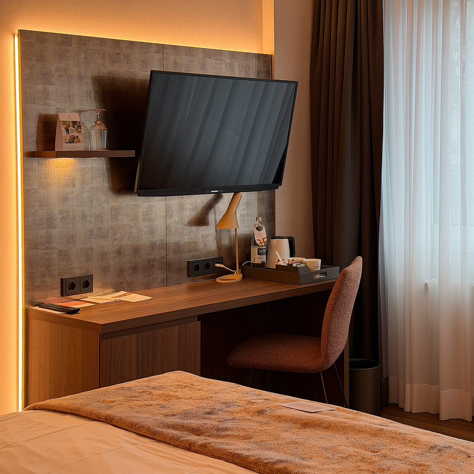 Ein modern eingerichtetes Hotelzimmer mit einem Schreibtisch und einem Fernseher an der Wand. Neben dem Schreibtisch steht ein Stuhl, und auf dem Tisch befinden sich ein Glas Wasser und einige Notizen. Eine große Fensterfront lässt Licht in den Raum.