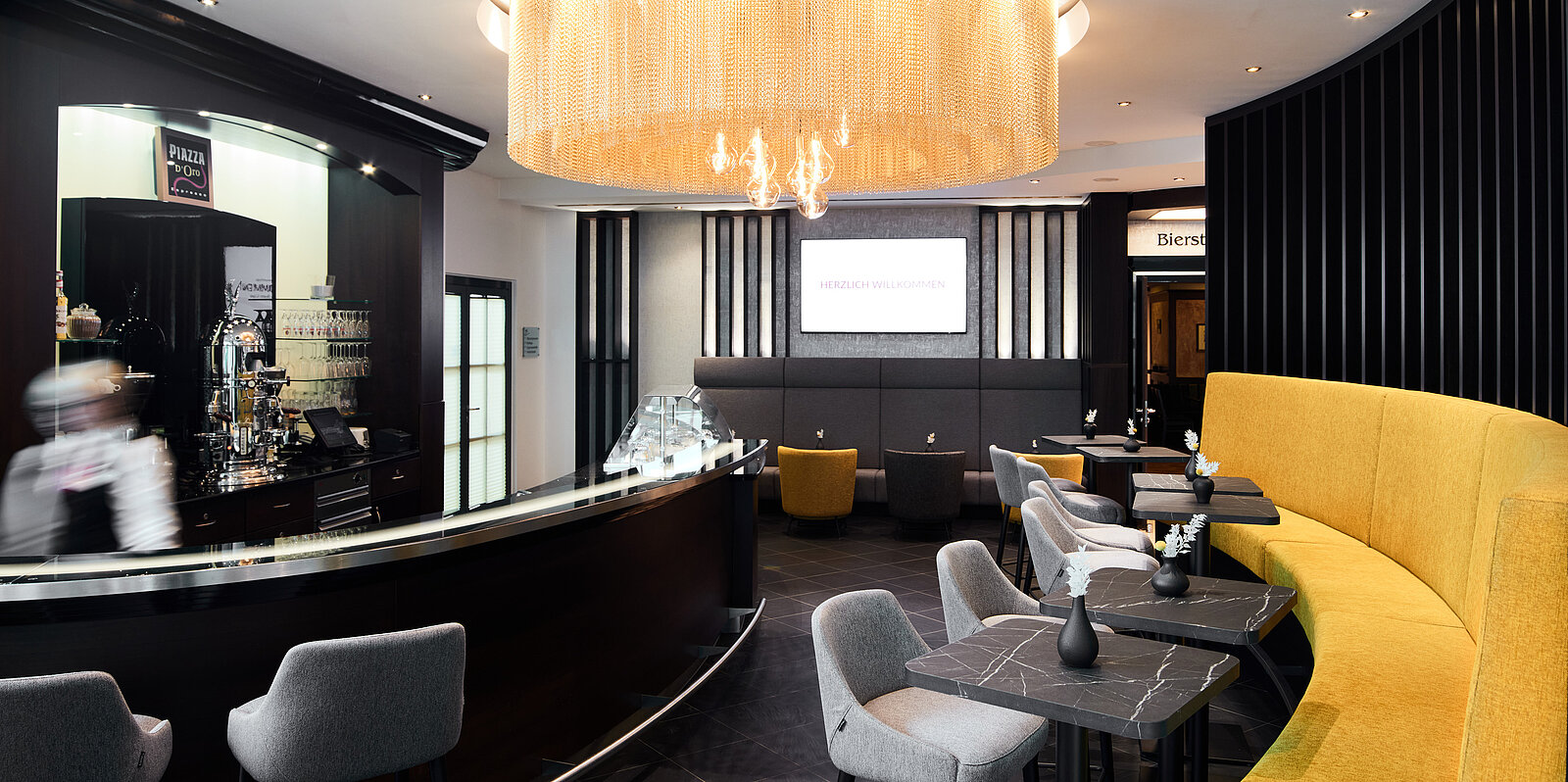 Eine elegante Cafénumgebung mit einem schwarz-weißen Interieur und hellen Sitzbereichen. Ein großer, moderner Kronleuchter hängt von der Decke. Die Bar ist gut ausgestattet, und der Raum strahlt eine einladende Atmosphäre aus.