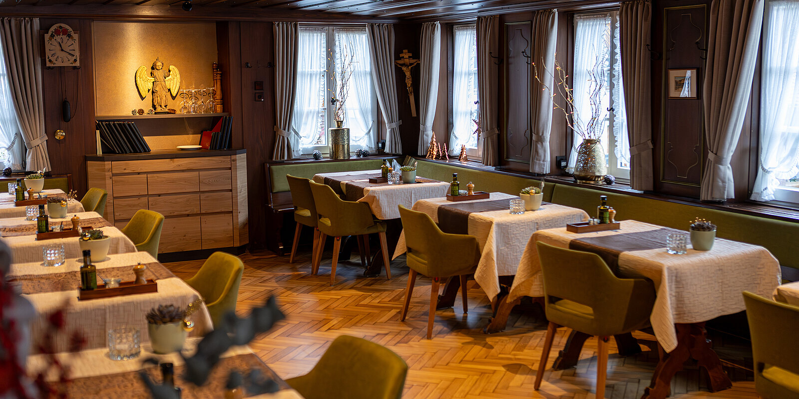 Gemütliches Restaurant mit Holzboden, Tischen mit weißen Tischdecken und grünen Stühlen. Die Fenster mit Vorhängen lassen viel Licht herein. Dekorationen und Pflanzen schaffen eine warme Atmosphäre.