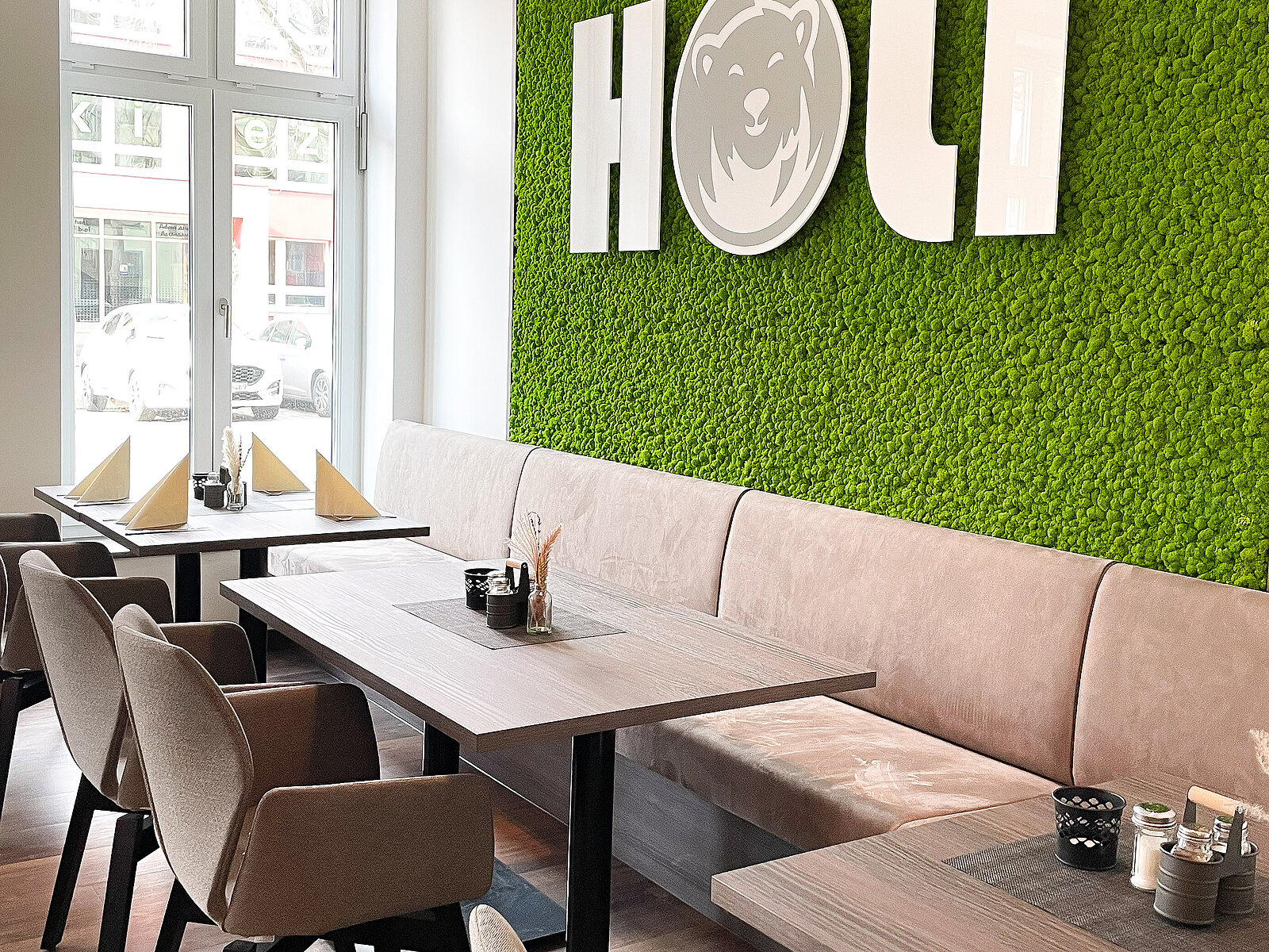 Gemütliches Café mit grüner Mooswand und großen weißen Buchstaben. Holz- und Polstermöbel bieten Sitzplätze für Gäste. Fenster im Hintergrund lassen Tageslicht herein.