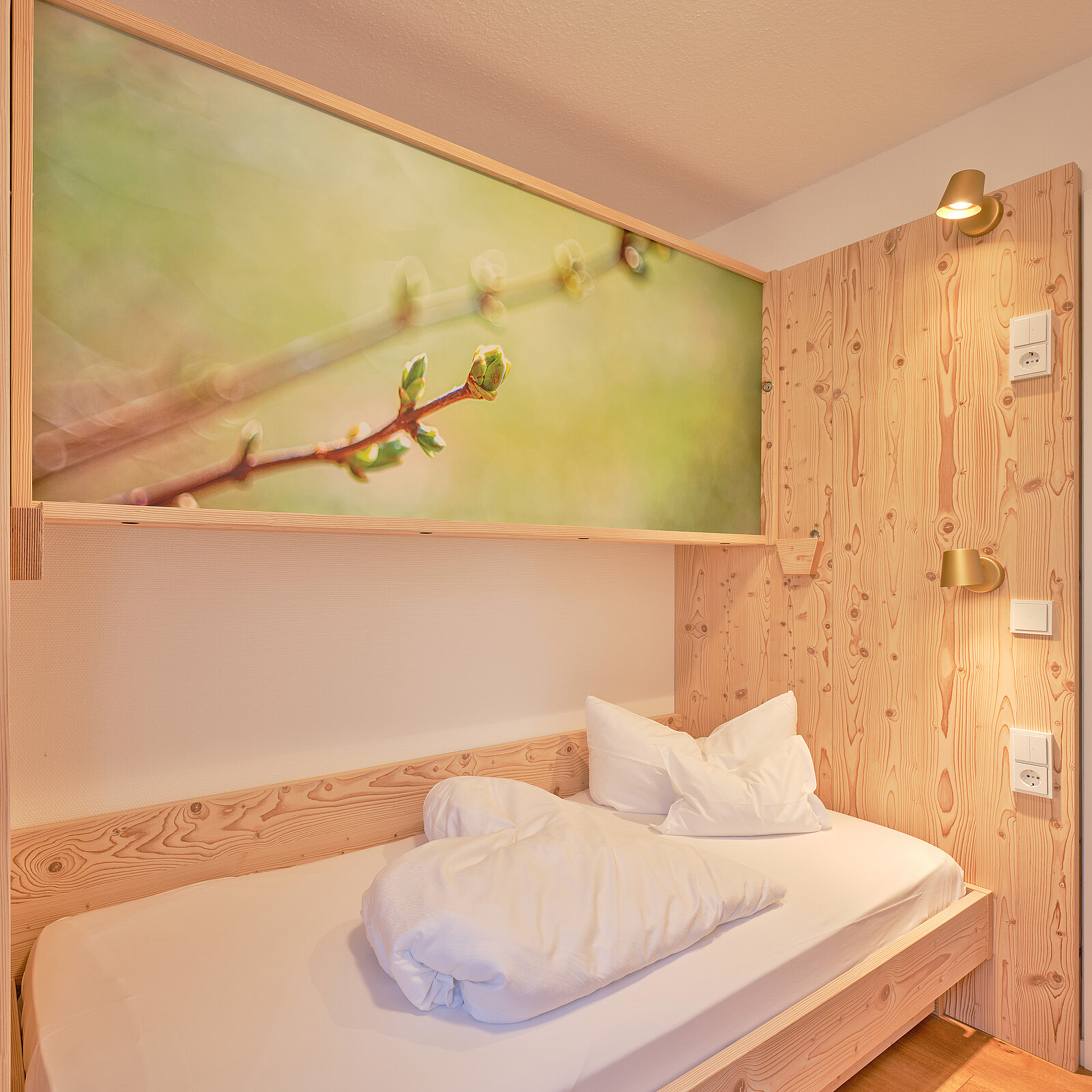 Ein schlicht eingerichtetes Einzelbettzimmer mit Holzwänden. Über dem Bett hängt ein großes Bild eines Zweigs mit Knospen. Die Bettwäsche ist weiß auf einem Holzrahmen. Eine Lampe ist an der Wand über dem Bett angebracht und sorgt für warmes Licht.