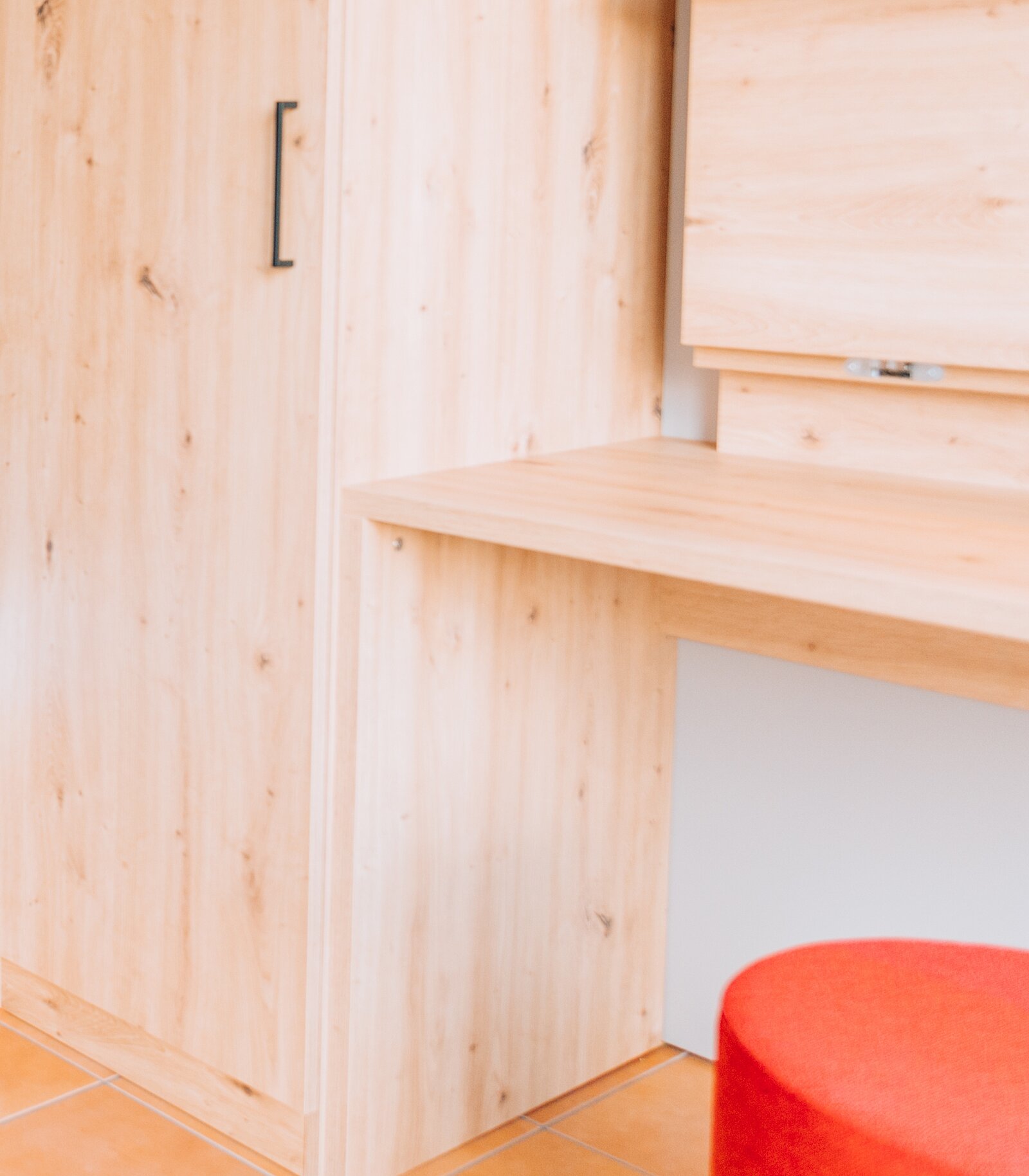 Ein Holzschreibtisch mit klaren Linien steht neben einem Holzschrank. Der Tisch zeigt eine abgeschrägte Kante. Rechts vom Tisch befindet sich ein runder, roter Hocker. Die gesamte Gestaltung vermittelt ein modernes und minimalistisches Ambiente.