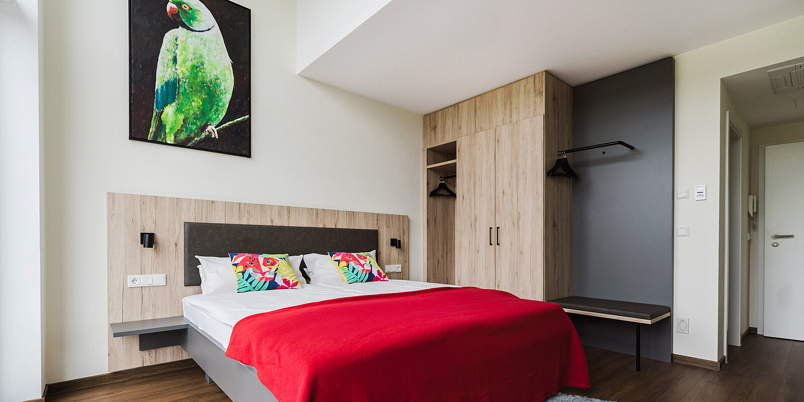 Modernes Schlafzimmer mit einem großen Bett mit roter Decke und bunten Kissen, einem Holzkleiderschrank und einem Bild eines Papageis an der Wand.