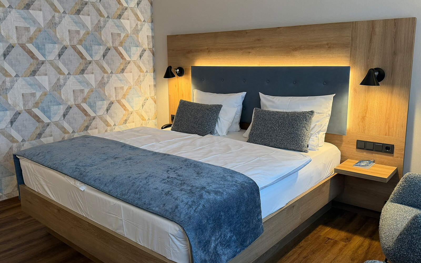 Das Bild zeigt ein modernes Schlafzimmer mit einem Doppelbett, das mit weißen Bettwäsche und blauen Kissen dekoriert ist. An der Wand hinter dem Bett befindet sich eine sanfte Beleuchtung. Neben dem Bett steht ein Nachtisch mit einer kleinen Lampe und einem Telefon. Die Wände sind mit einem geometrischen Muster dekoriert.