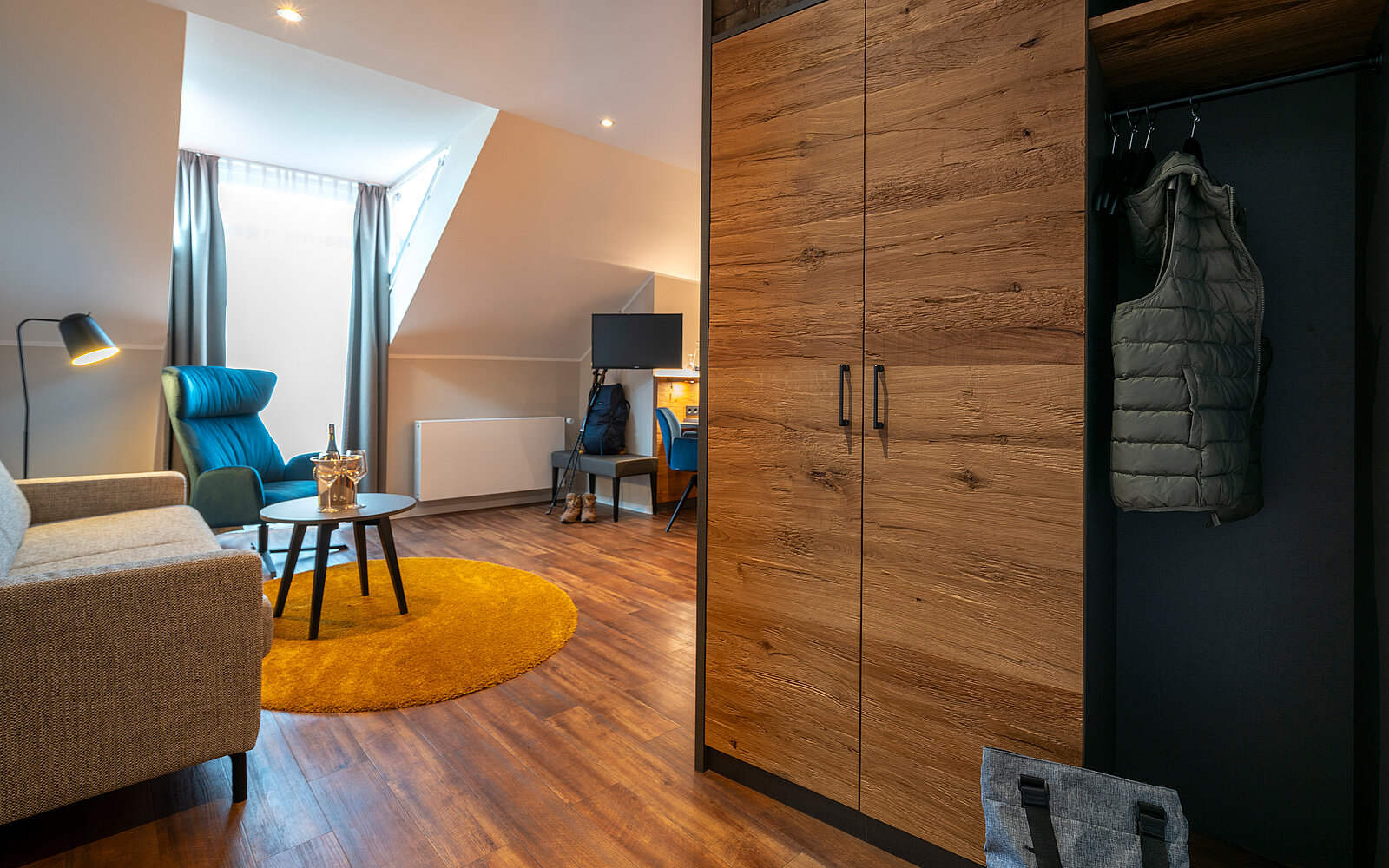 Ein modernes Zimmer mit Holzboden. Links steht ein gemütliches Sofa neben einem runden Tisch auf einem gelben Teppich. Rechts befindet sich ein Schrank mit einer grauen Weste, und im Hintergrund ist ein Bildschirm auf einem Tisch zu sehen, daneben ein blauer Sessel und ein Fenster mit Vorhängen.