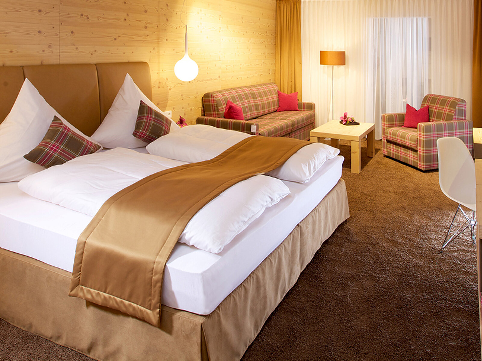 Ein gemütliches Hotelzimmer mit einem großen Doppelbett, das mit einer goldenen Bettdecke bedeckt ist. Auf dem Bett liegen zwei Kissen mit kariertem Muster. Im Hintergrund stehen ein Sofa und zwei Sessel, ebenfalls mit kariertem Stoff. Die Wände sind aus Holz, und es gibt eine helle Beleuchtung.