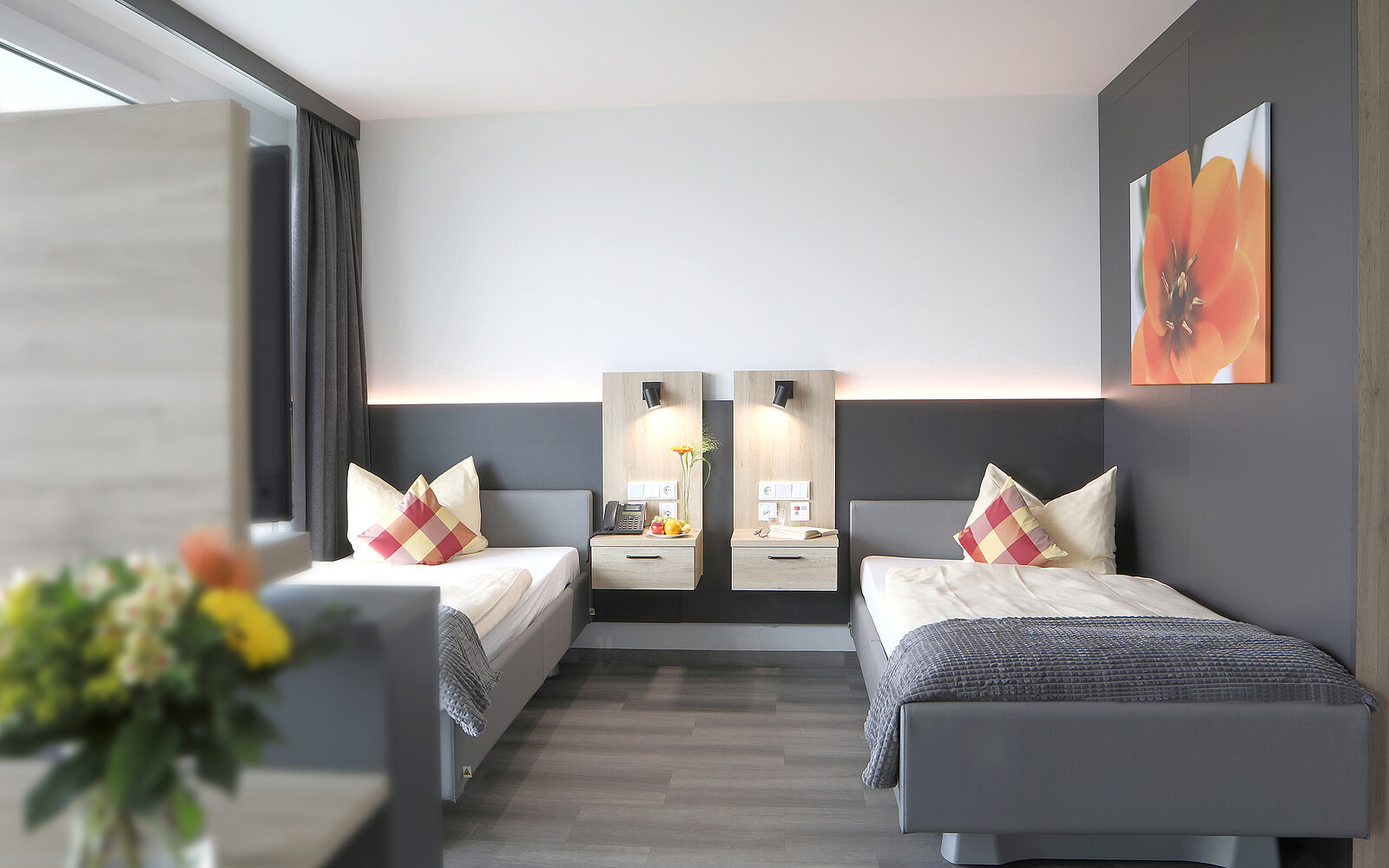 Ein modernes Zimmer mit zwei Einzelbetten, weißen Bettdecken und dekorativen Kissen. An der Wand befinden sich Nachttische mit Lampen und Steckdosen. Eine große, farbige Wanddekoration und ein Blumenstrauß im Vordergrund ergänzen die Einrichtung. Der Raum wirkt hell und einladend.