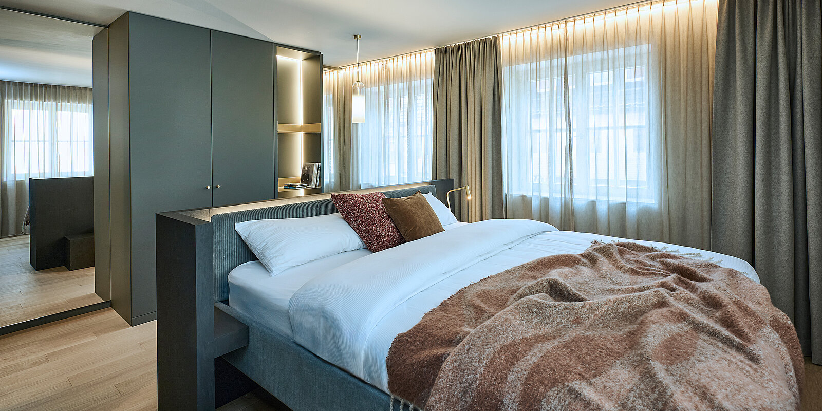 Ein modernes Schlafzimmer mit großem Bett, weißen Laken und dekorativen Kissen. Links steht ein grauer Kleiderschrank. Tageslicht strömt durch große Fenster mit beigen Vorhängen. Eine warme Decke liegt über der Betthälfte. Holzfußboden rundet das elegante Ambiente ab.
