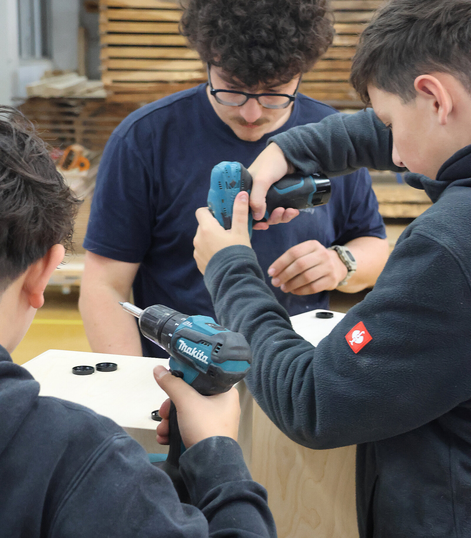 Drei Personen arbeiten gemeinsam an einem Holzprojekt in einer Werkstatt. Zwei junge Personen halten elektrische Schraubendreher, während ein Erwachsener sie anleitet. Im Hintergrund sind Holzregale sichtbar.