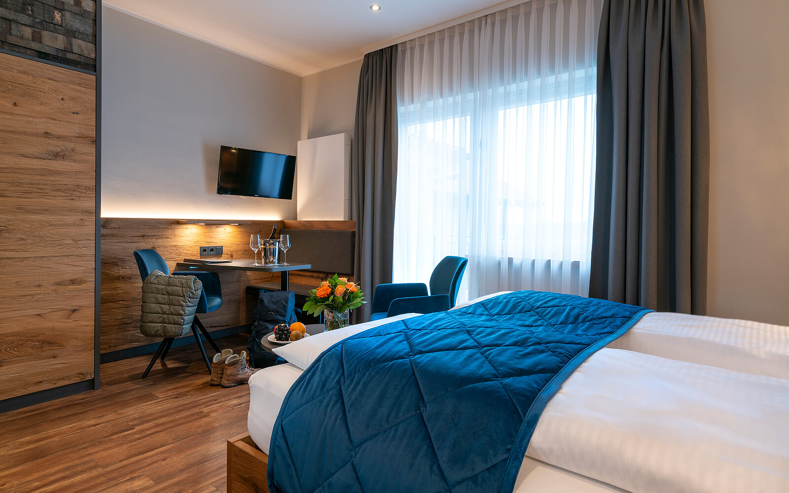 Ein modernes Hotelzimmer mit einem Doppelbett und einer blauen Bettdecke. Neben dem Bett steht ein Tisch mit zwei Stühlen, einer Blumenvase und einem Teller mit Obst. Große Fenster lassen viel Tageslicht herein und es gibt einen Fernseher an der Wand.