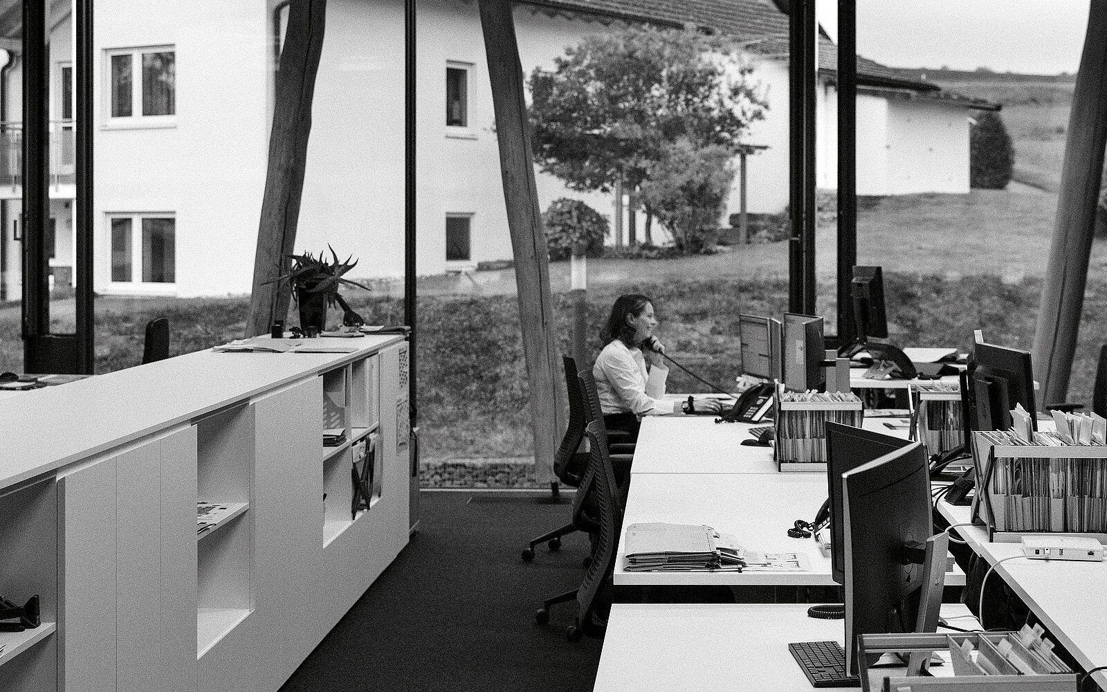 Eine Frau sitzt in einem modernen Büro an einem Schreibtisch und telefoniert. Große Fenster zeigen einen Blick auf draußen.