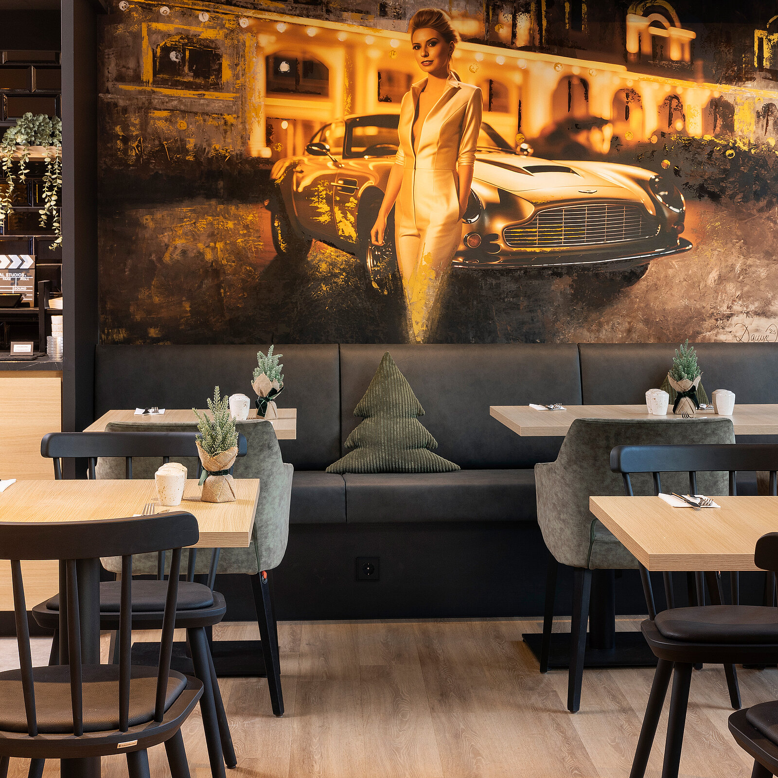 The image features a stylish interior of a restaurant, showcasing a wall mural of a glamorous woman in a white outfit next to a classic car. Tables and chairs are arranged in the foreground, with small plants adding a touch of greenery. The ambiance is modern and sophisticated.