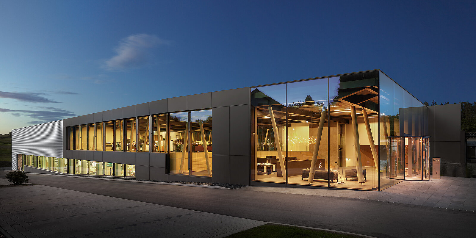 Das Bild zeigt ein modernes Gebäude mit großen Glasfenstern und einem eleganten Design, das am Abend beleuchtet ist. Die Struktur weist eckige Elemente auf und steht vor einem dämmerigen Himmel, wodurch ihre zeitgenössische Architektur hervorgehoben wird. In der Nähe befindet sich ein gepflasterter Bereich, der zum Eingang führt.