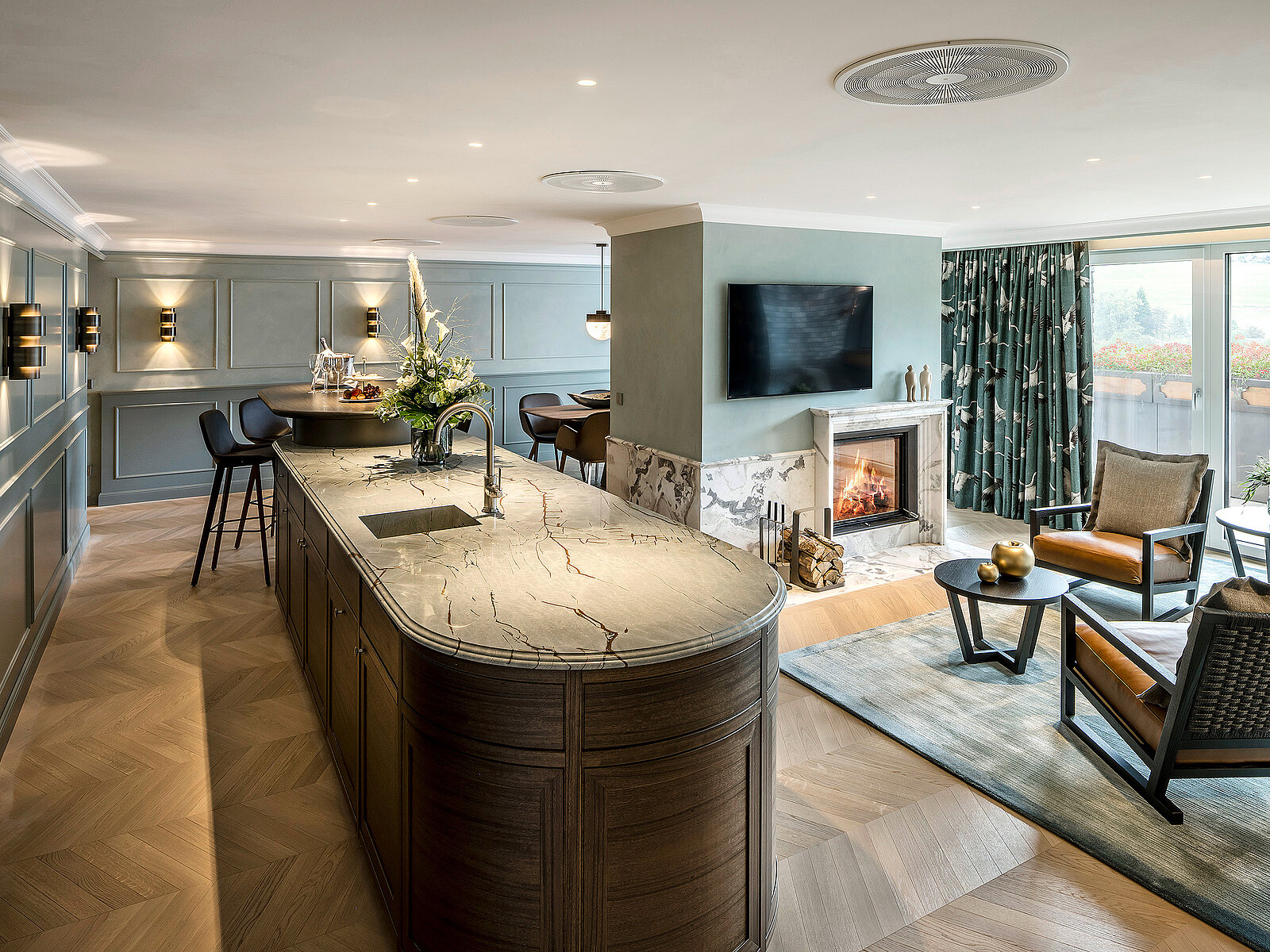 In einer eleganten Küche mit einer Insel aus Stein, einem modernen Fernseher an der Wand und einem Kamin. Der Raum ist hell, mit Holzfußboden und stilvollen Möbeln, darunter ein gemütlicher Sessel und ein Tisch mit Dekoration. Große Fenster bieten Ausblick auf die Außenlandschaft.