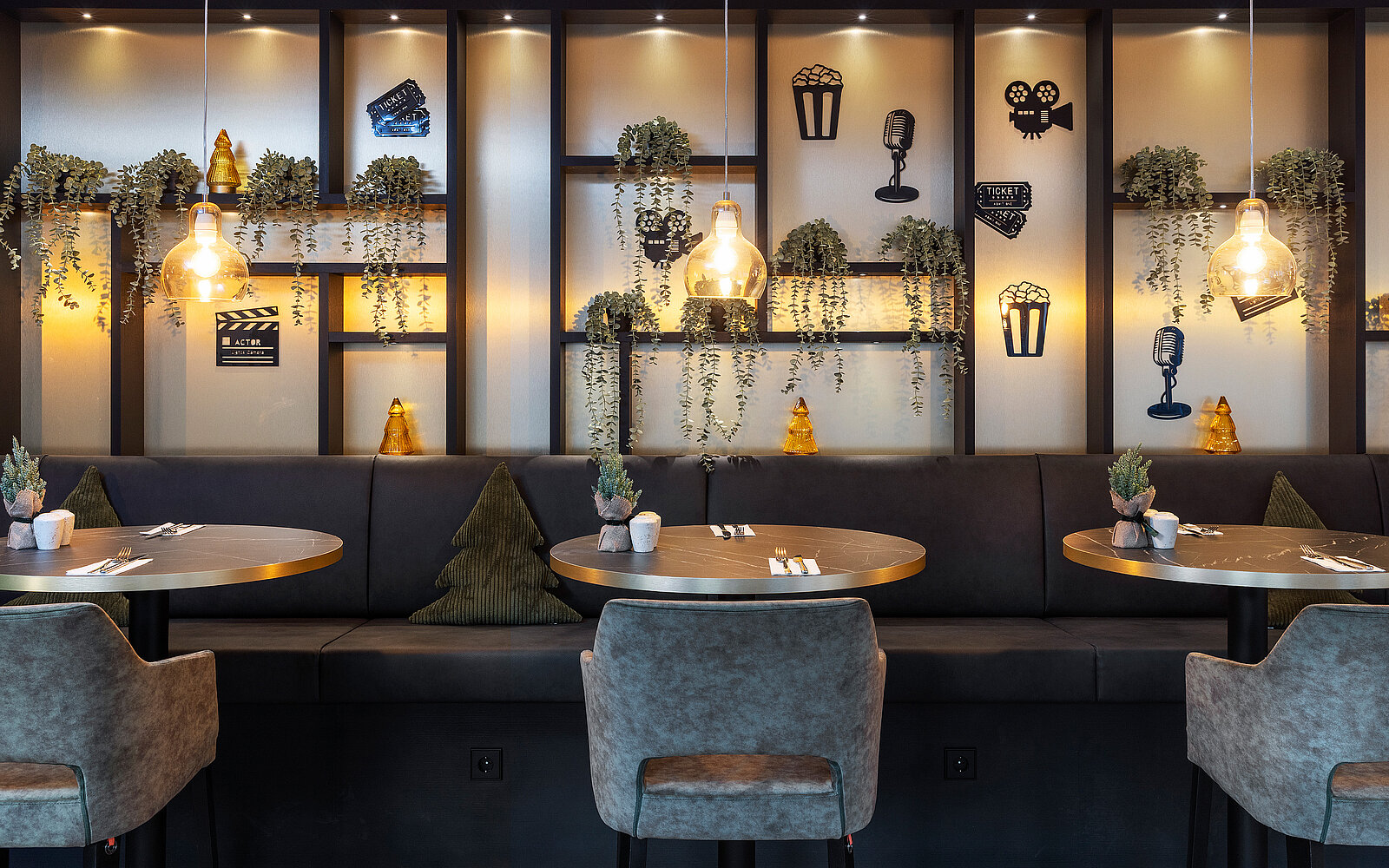 The image depicts a cozy restaurant interior featuring two round tables with chairs. A dark wall behind them is adorned with shelves displaying decorative items and greenery. Soft lighting creates a warm atmosphere, enhancing the inviting dining space.