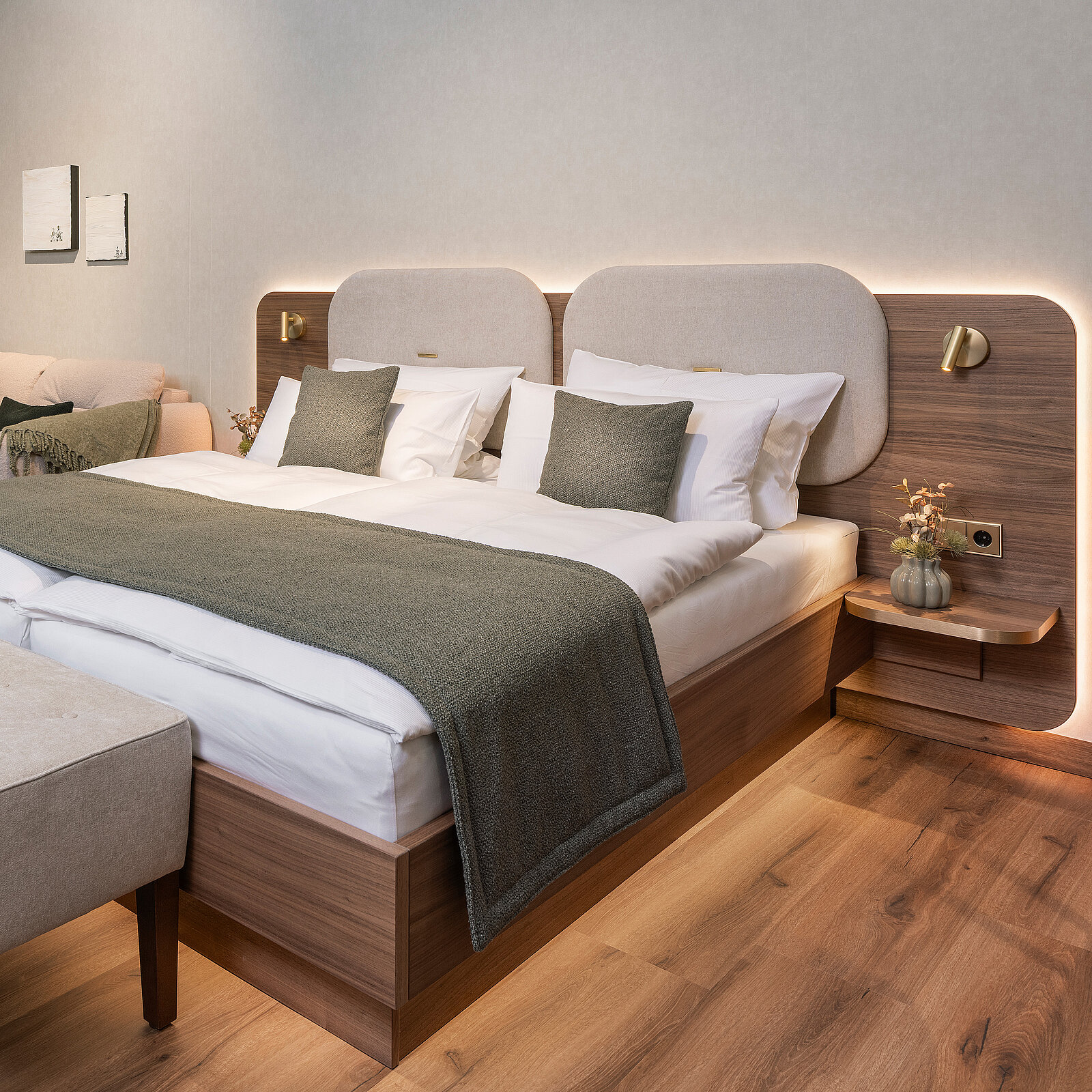 A modern hotel room featuring a spacious bed with plush white linens and gray accent pillows. The headboard is softly illuminated, enhancing the cozy ambiance. A small bedside table holds a decorative vase. Light wood flooring adds warmth to the room's contemporary design.
