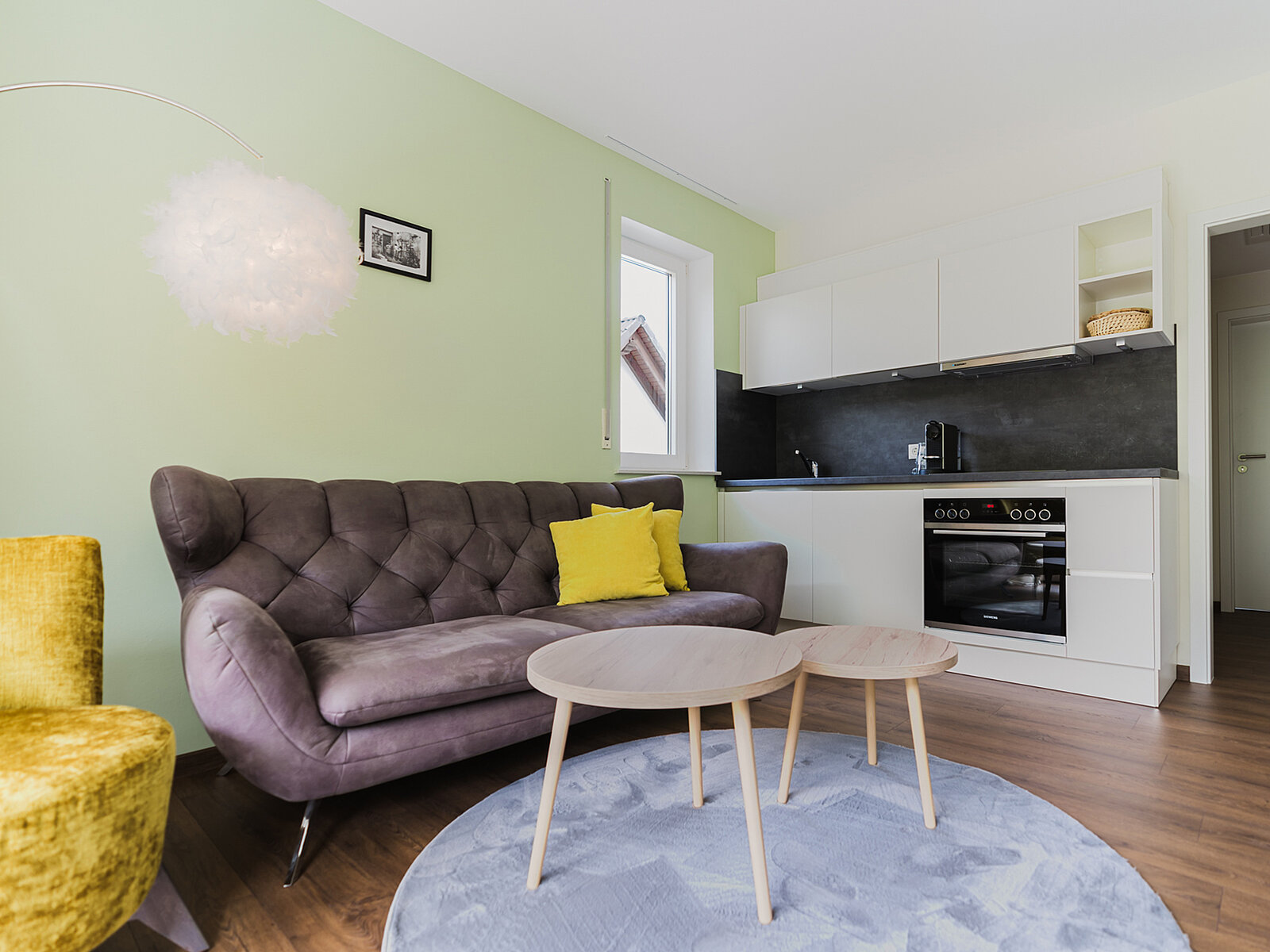 Ein modernes Wohnzimmer mit einer grünen Wand, einem grauen Sofa mit gelbem Kissen und einem runden, blauen Teppich. Zwei helle Holztische stehen vor dem Sofa. Im Hintergrund ist eine kleine, weiße Küche mit schwarzen Arbeitsflächen zu sehen.
