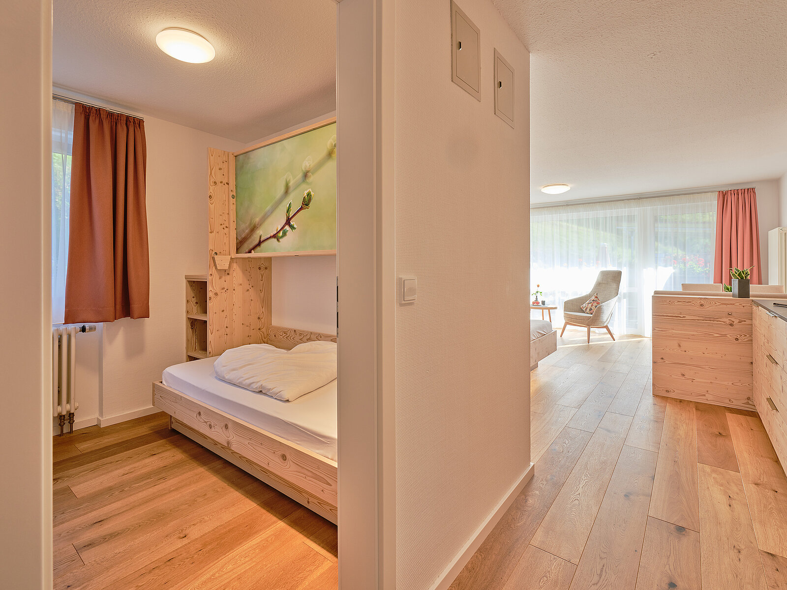 Gemütliches Interieur eines modernen Apartments. Links ein Schlafzimmer mit Holzbetten und dezenten Vorhängen, rechts ein heller Wohnbereich mit großen Fenstern, Holzfußboden und eleganter Einrichtung. Natürliche Materialien und sanfte Farben schaffen eine einladende Atmosphäre.